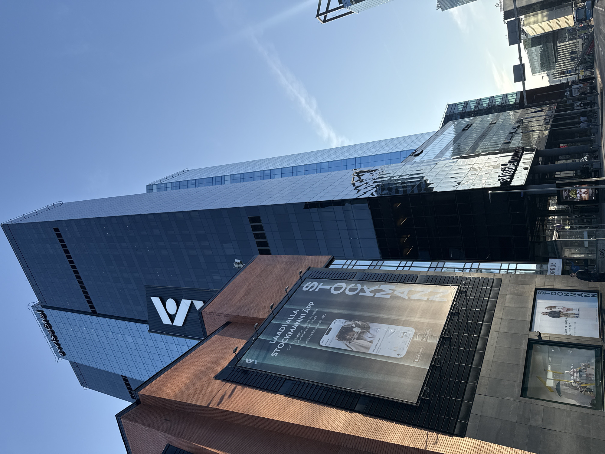 The image shows a street scene in Tallinn, Estonia, focusing on the Swissôtel Tallinn and the adjacent Stockmann department store. The Swissôtel is a tall, modern skyscraper with a glass facade, reflecting the blue sky. Above the hotel's entrance, the "Swissôtel" logo is visible. To the left of the hotel, a brick building with a large billboard advertises the "STOCKMANN APP." The billboard displays a smartphone screen with the Stockmann logo and an image of a person. Further down the street, other buildings and traffic are visible, including streetlights and a bus. The sky is clear and blue with a faint cloud streak. Several individuals are present on the sidewalk, some walking and others standing. The overall scene depicts an urban environment with a focus on commercial and hospitality buildings.
