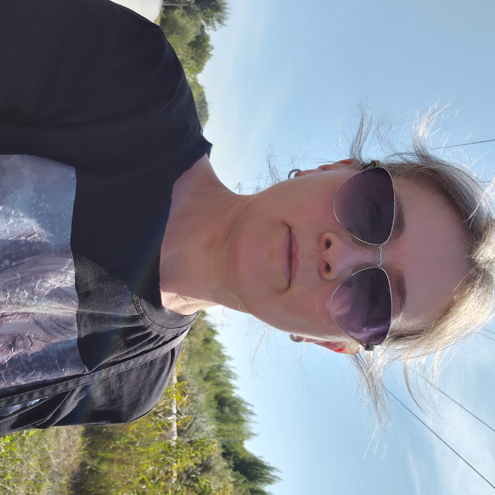 A medium shot captures a woman outdoors, likely in a rural or semi-rural area, on a sunny day. She is looking directly at the camera. She has blonde hair, which is slightly windswept, and is wearing large, dark sunglasses with a metallic frame. She has small hoop earrings in her ears. She is wearing a black t-shirt with a graphic design on the lower portion. A strap from a bag or backpack is visible on her right shoulder. The background consists of a clear blue sky with some power lines, and a grassy area with trees and bushes. Based on the style of the power lines and the vegetation, this scene could be located in a suburban or rural area on the outskirts of Novosibirsk, Russia.