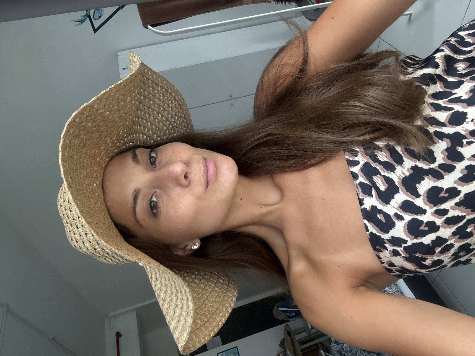 A young woman is featured in a close-up, rotated selfie, likely taken indoors in Kuala Lumpur, Malaysia. She has long, straight brown hair, light brown eyes, and a subtle, pleasant smile. Her skin is fair, showing visible freckles. She is wearing a wide-brimmed, light-brown straw hat and a strapless top or dress with a distinct black and brown leopard print pattern on a cream background. A small, round silver stud earring is visible in her right ear.
The background is simple, featuring a light gray or white wall. Above her, a white horizontal railing or shelf is visible, along with a dark brown object, possibly part of a furniture piece or a hanging item. In the lower right corner, a small framed picture with blue and white detailing can be partially seen. The lighting is soft and natural, indicating it is daytime. The image captures a candid, personal moment. No text is visible.