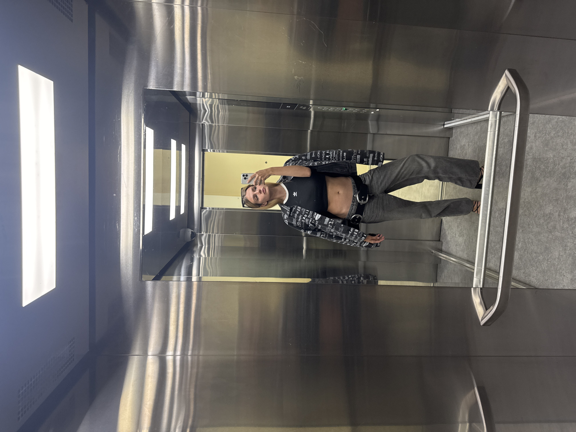 The image shows a person taking a selfie in an elevator. The elevator's interior is stainless steel with a rectangular light panel on the left. There is a mirror on the back wall, reflecting the person and the elevator's interior. The person is wearing a black crop top, jeans, and a patterned jacket. They are holding a phone in their right hand. There is a handrail attached to the right wall. The elevator floor is gray. The location is Unknown, Unknown.