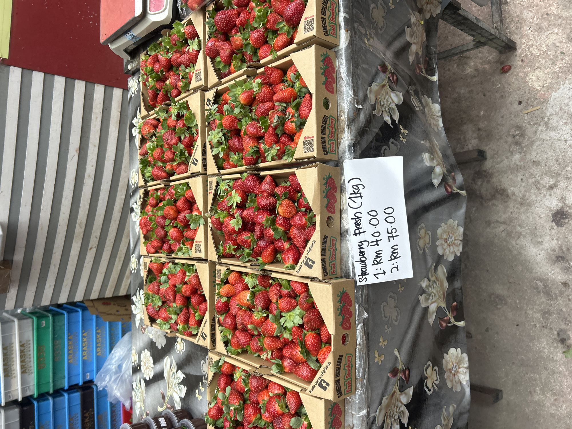 In the city of Tanah Rata, Malaysia, a market stall displays an abundance of fresh strawberries. The strawberries are neatly arranged in cardboard boxes, each filled to the brim with ripe, red fruit and green leaves. The boxes are labeled with "Cameron Highlands Fresh Strawberry" and feature a Facebook logo and a QR code. A white sign is placed in front of the strawberries, indicating prices for the fruit: "Strawberry Fresh (1kg) 1: Rm 40.00 2: Rm 75.00". Behind the stall, stacks of blue and green plastic crates are visible, some bearing the label "ARASKА". A red and white weighing scale sits on the counter to the right. The stall itself is covered with a dark tablecloth adorned with a floral pattern. The ground is made of concrete, with a single fallen strawberry and a small wooden stick visible on it.