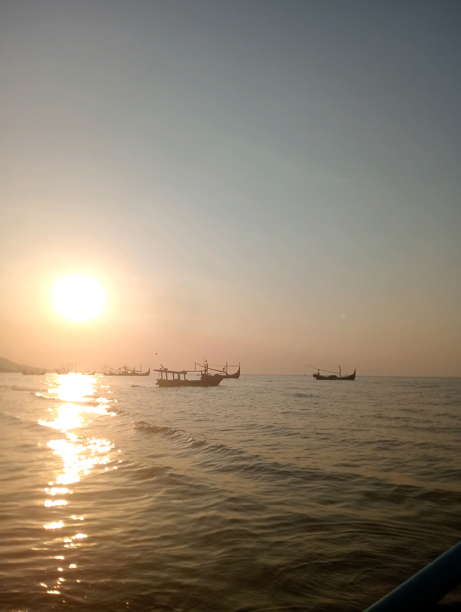 A vertical, wide-angle shot captures a serene ocean scene at sunset in Pesisir, Indonesia. The sun, a bright, diffused orb, hangs low in the left portion of the sky, casting a shimmering golden path across the gentle waves. Several traditional Indonesian fishing boats, silhouetted against the horizon, are scattered across the water, some with sails or masts visible. The sky transitions from a pale yellow near the horizon to a soft blue at the top. The water is calm with small ripples, reflecting the warm light of the setting sun. In the foreground, the edge of a blue object, possibly part of the boat from which the photo is taken, is visible on the right. The overall atmosphere is peaceful and tranquil.