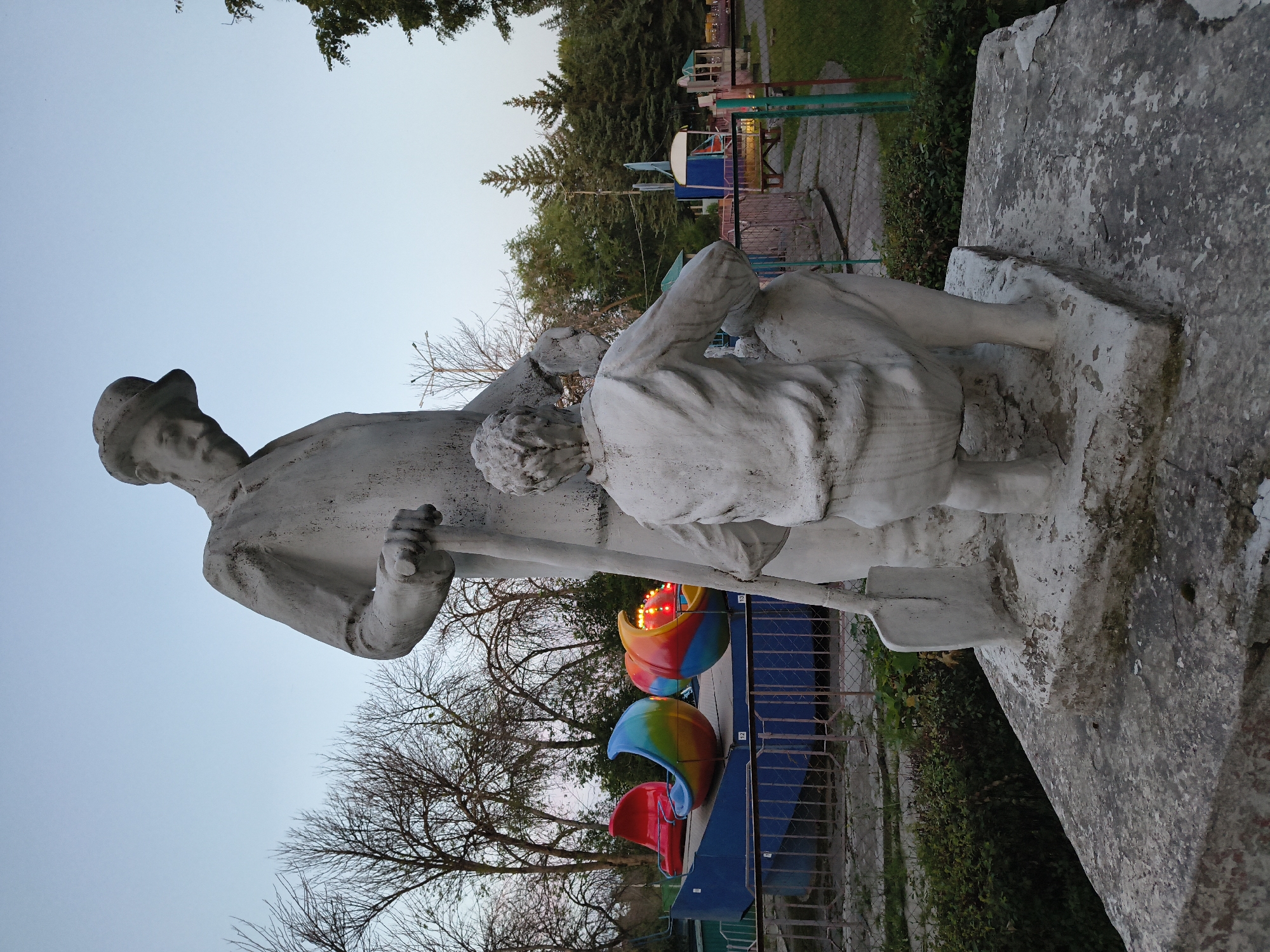 A white concrete sculpture depicts two figures, one standing and one kneeling, in an outdoor setting. The standing figure, wearing a hat and coat, holds a shovel. The kneeling figure is facing away from the viewer. In the background, there is a colorful amusement park ride with multiple seats, fences, trees, and a clear sky. The sculpture is situated on a stone pedestal. The overall scene suggests a park or recreational area in Mikhaylovka, Russia.