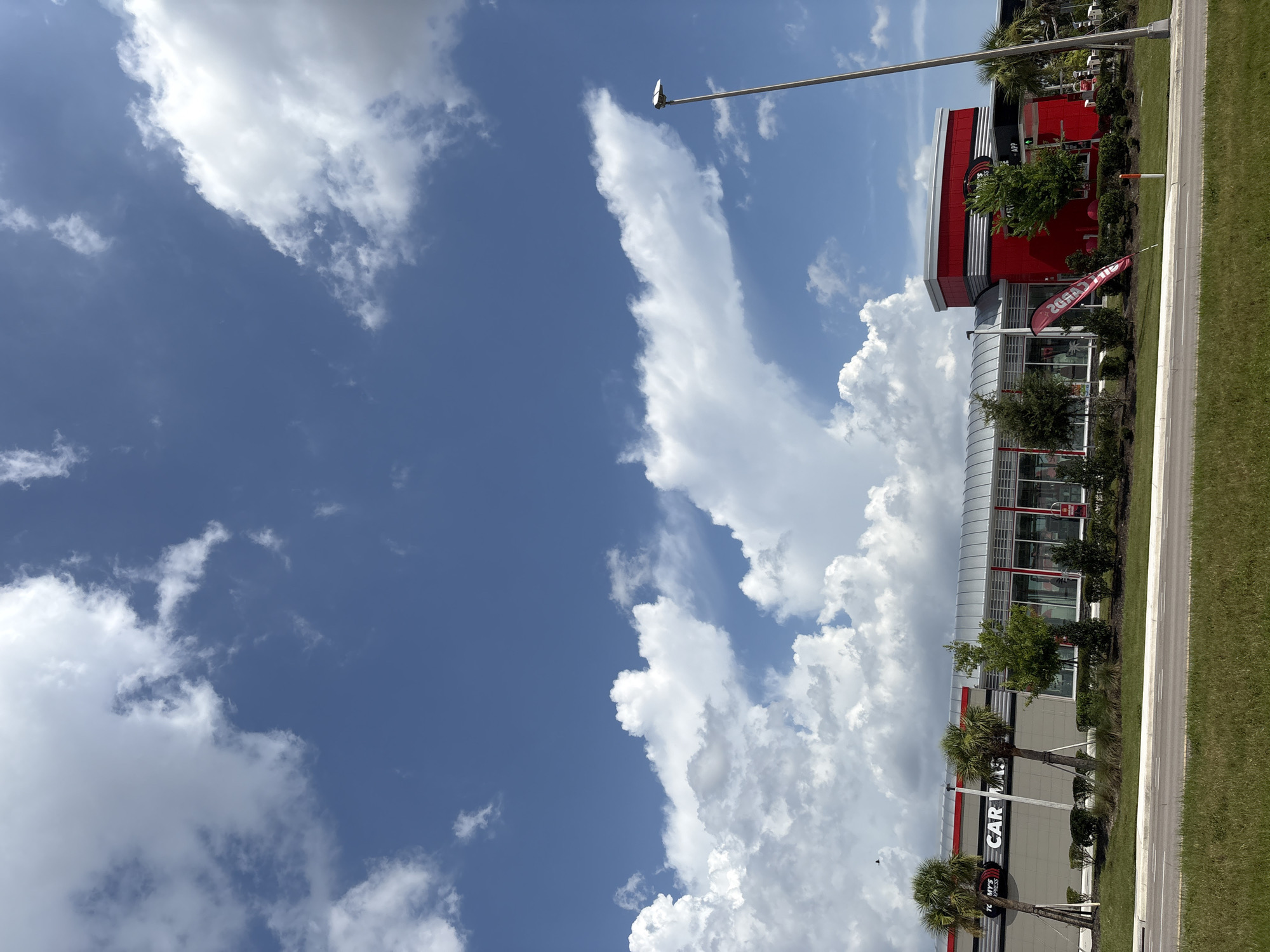 In Parrish, Florida, United States, a Tommy's Express Car Wash is captured under a bright, partly cloudy sky. The car wash features a modern architectural design with a long, silver, curved roofline and prominent red accents. The building has large glass windows and doors, suggesting an open and accessible space. In front of the building, lush green grass and well-maintained landscaping, including palm trees and various shrubs, line the road. A tall streetlight stands to the right of the building. The sky is a vibrant blue, dotted with large, fluffy white cumulus clouds that create dynamic shadows and highlights across the scene. One red flag with white text is planted in front of the building, and the words "CAR WASH" are visible on the building's facade.