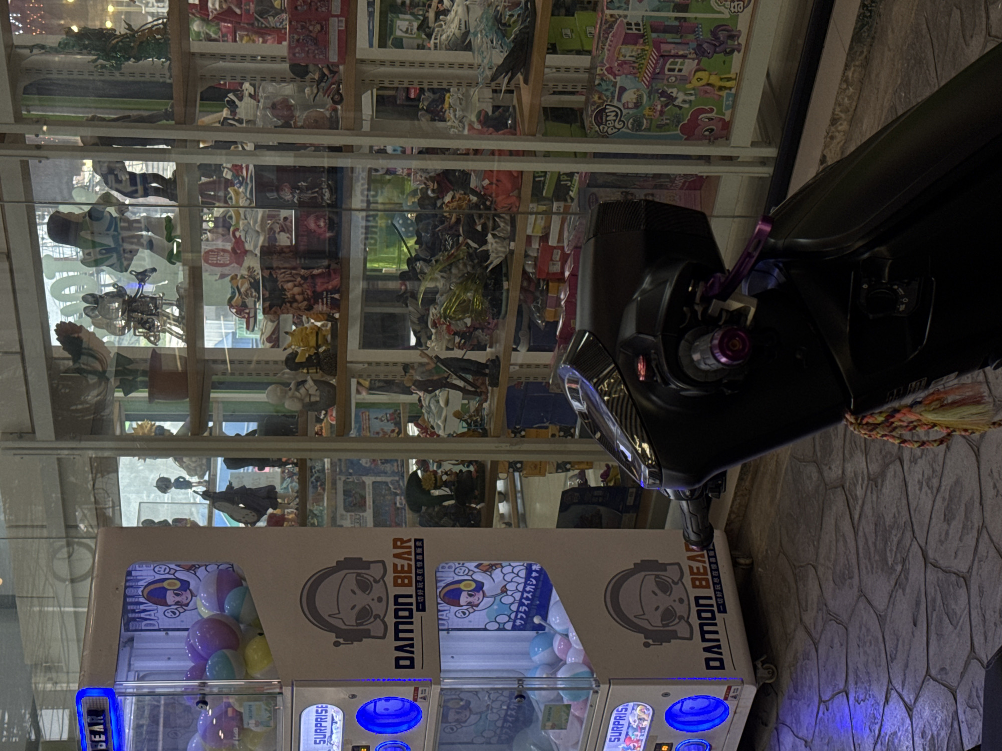 The image depicts a street-level view in Karon, Thailand, focusing on a storefront and a scooter. To the left, a white, illuminated vending machine with blue lights displays the brand "DAMON BEAR" and is filled with colorful prize balls. Behind the vending machine, shelves are visible, lined with various collectible items and figurines, including what appear to be anime characters, sneakers, and other merchandise. To the right, a dark-colored scooter is parked, its handlebars and front section prominently featured in the foreground. A decorative tassel hangs from the scooter. The overall environment appears to be an indoor retail space or a covered outdoor area with a tiled floor.