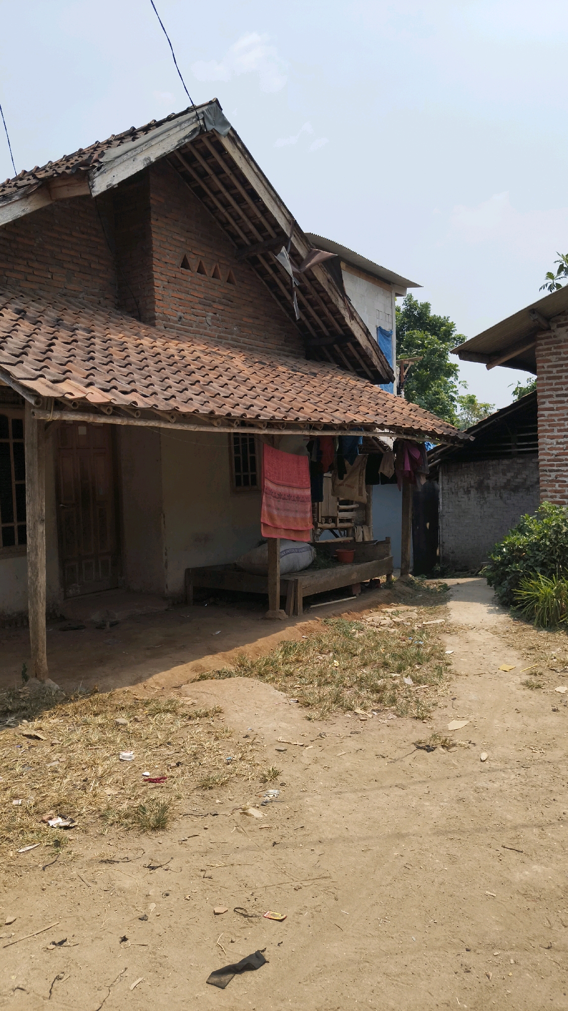 A medium shot captures a humble, rustic dwelling in Tenjo, Indonesia, under a clear, bright sky. The house, constructed with reddish-brown clay tiles on its roof and exposed brick walls, exhibits signs of wear and tear. A simple wooden porch extends from the front, supported by rough-hewn wooden posts. Clothes hang from a line strung across the porch, including a vibrantly colored red cloth. In front of the house, a low wooden bench serves as seating, with a white sack and a red plastic container visible on it. The yard in front of the house is unpaved, with patches of dry grass and scattered debris, suggesting a rural or semi-rural environment. To the right, a narrow dirt path leads away from the house, bordered by more dry grass and a dense green bush. Another structure, partially visible to the right, appears to be made of brick with a corrugated metal roof. The overall scene conveys a sense of everyday life in a modest Indonesian community.