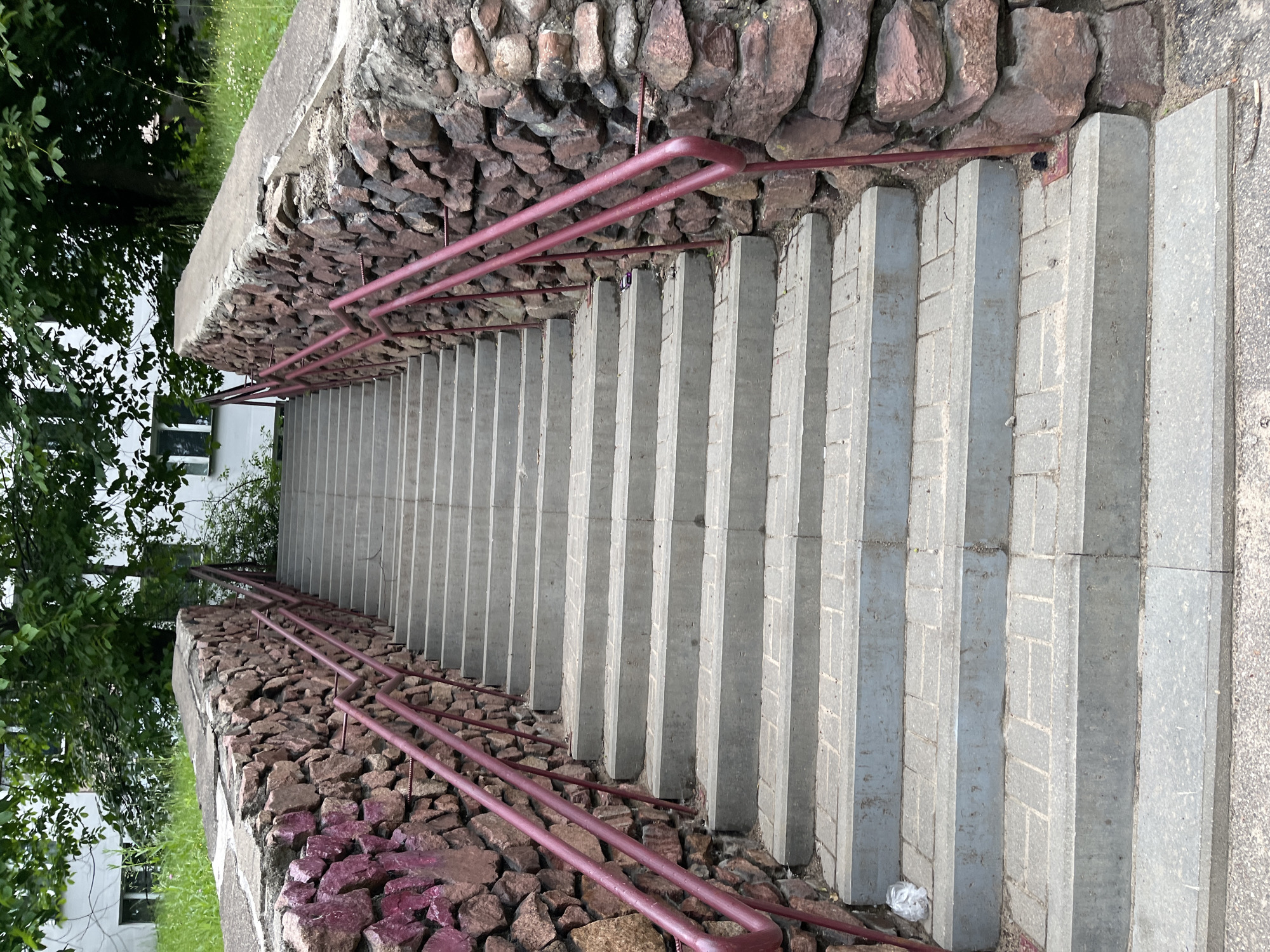 The image shows an outdoor stone staircase with a reddish-brown stone retaining wall on either side. The stairs are made of grey concrete with brick patterns on the risers. Red metal handrails run along both sides of the staircase. Green grass and trees are visible at the top of the stairs, and a white building with windows can be seen in the background. The scene appears to be in a public outdoor area.
