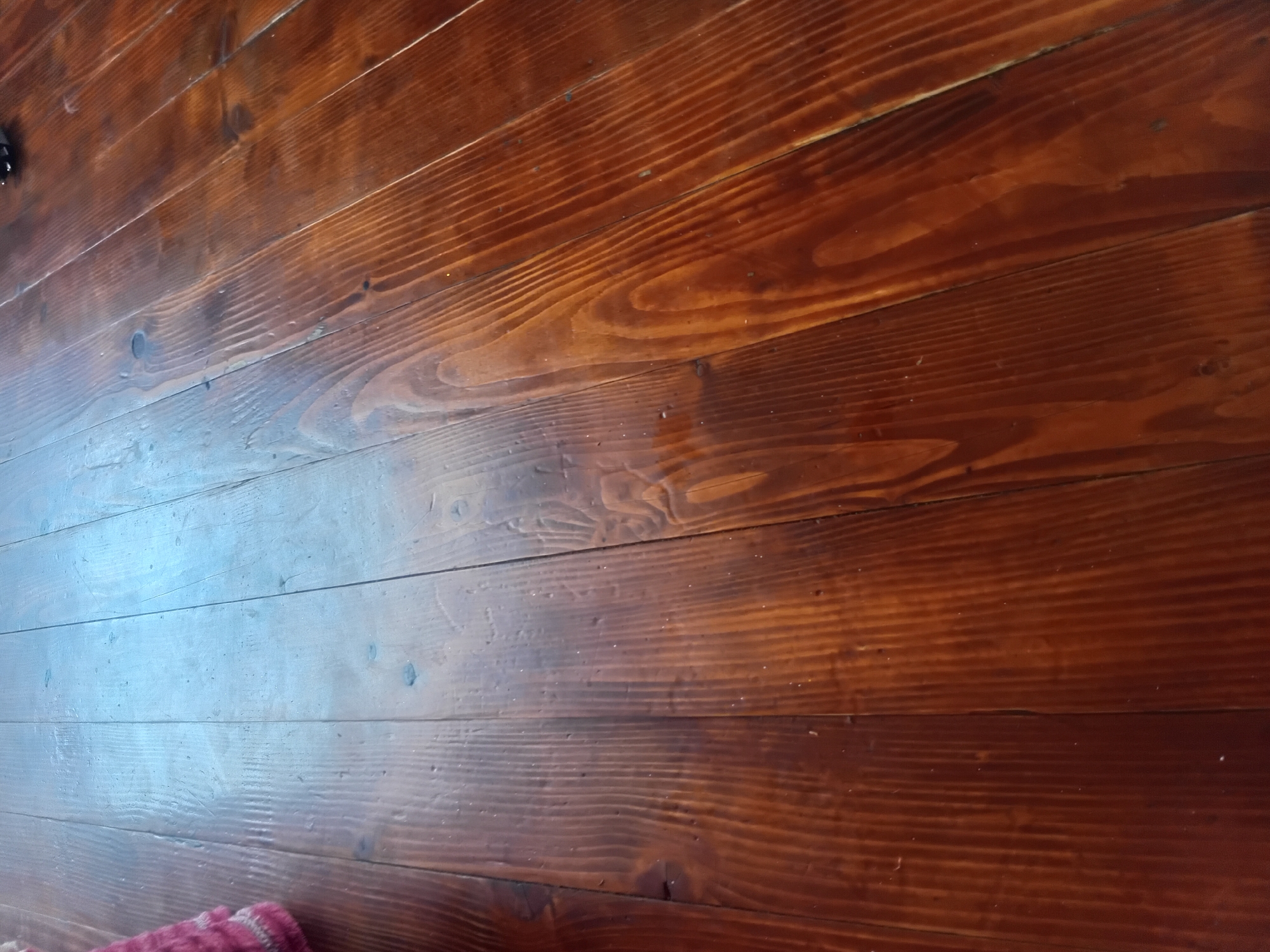 The image shows a close-up, overhead view of a polished wooden floor composed of narrow planks. The wood is a rich, dark brown color with visible grain patterns. Some planks show knots. Light reflects off the polished surface, creating bright highlights and darker shadows that emphasize the texture and direction of the wood grain. The planks are laid parallel to each other, running diagonally across the frame from the bottom left to the top right. In the upper left corner, a small portion of a pink and white knitted item is visible, suggesting it is resting on the floor. The scene appears to be indoors, given the flooring. There is no clear indication of the specific location within Verkhniachka, Ukraine, other than the flooring itself.