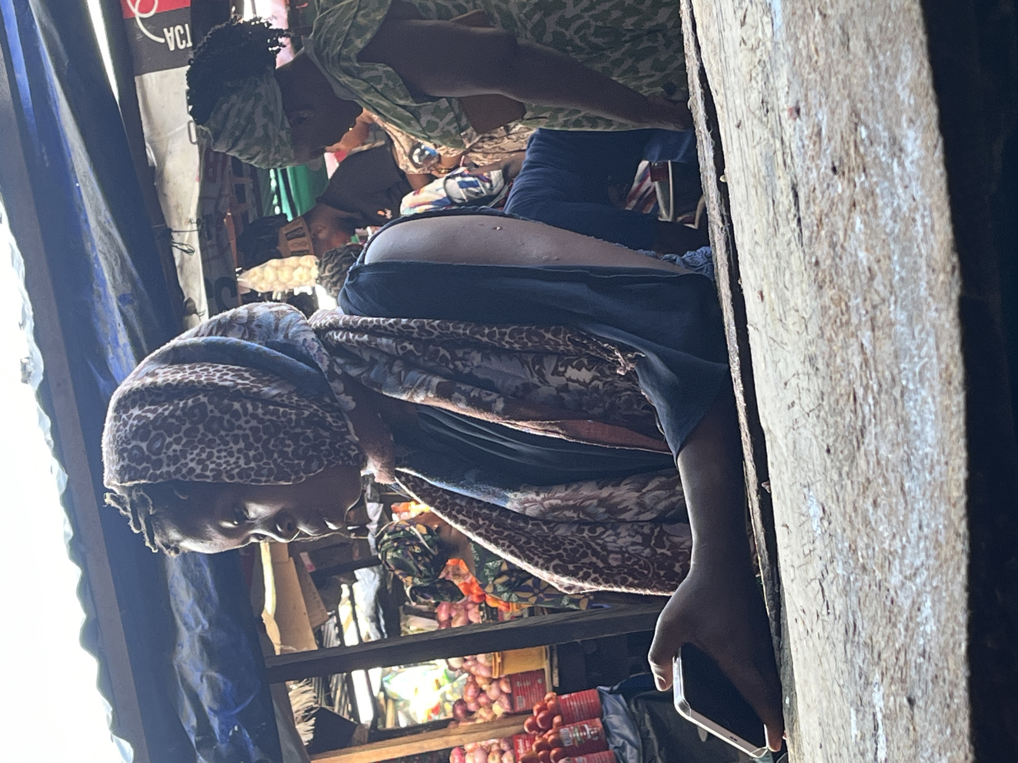 In Ado, Nigeria, the image captures a bustling market scene. A woman wearing a leopard-print headscarf and patterned shawl is looking down and holding a smartphone in her right hand, close to a wooden structure, possibly a stall frame. Behind her, numerous vendors and customers are visible, some with patterned headscarves. Produce, including onions and tomatoes, is displayed on shelving in the background. The setting appears to be a covered market area, given the presence of tarpaulin or fabric awnings overhead.