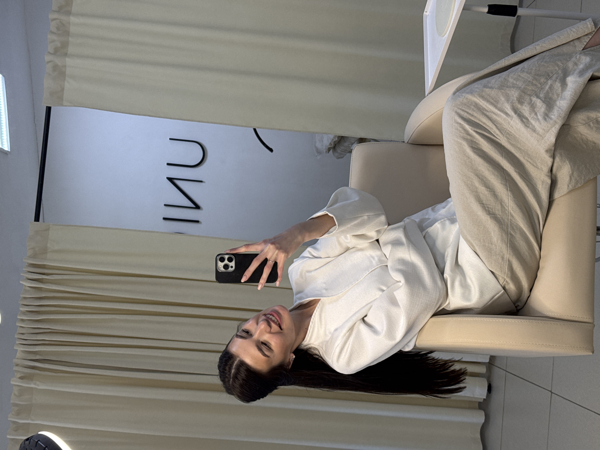 A woman in a white robe and beige pants sits in a beige chair in what appears to be a clinic in Chaikovsky, Russia. She is holding a black smartphone, presumably taking a selfie. The background includes curtains, a logo on the wall, and part of a table. The environment is well-lit.