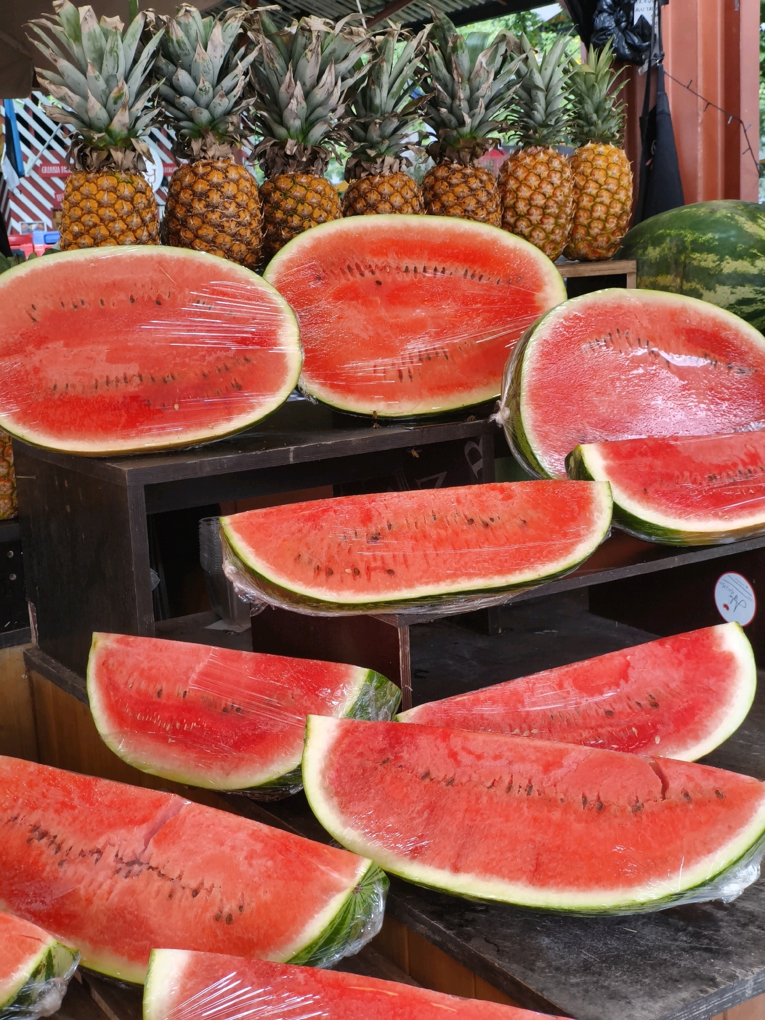 A close-up shot captures a vibrant display of fruits, prominently featuring several watermelons sliced in half and wrapped in plastic, alongside a row of ripe pineapples. The watermelons are arranged on dark wooden shelves and a table, showcasing their bright red flesh and green rinds. Some are whole, while others are cut into wedges, some still retaining their rind. A large, whole green watermelon is visible in the background, to the right of the pineapples. The pineapples are golden yellow, with spiky green crowns, and are neatly lined up behind the watermelons. The setting appears to be an outdoor market or a fruit stand, with a hint of a rustic wooden structure in the background and what looks like part of a black tarp or bag. The lighting suggests it might be daytime, possibly outdoors, given the natural appearance of the fruits. There are no people visible in the frame. The scene is a still-life composition focused on the fresh produce, suggesting a focus on healthy eating and local markets. The image does not contain any elements that would place it specifically in Tallinn, Estonia.