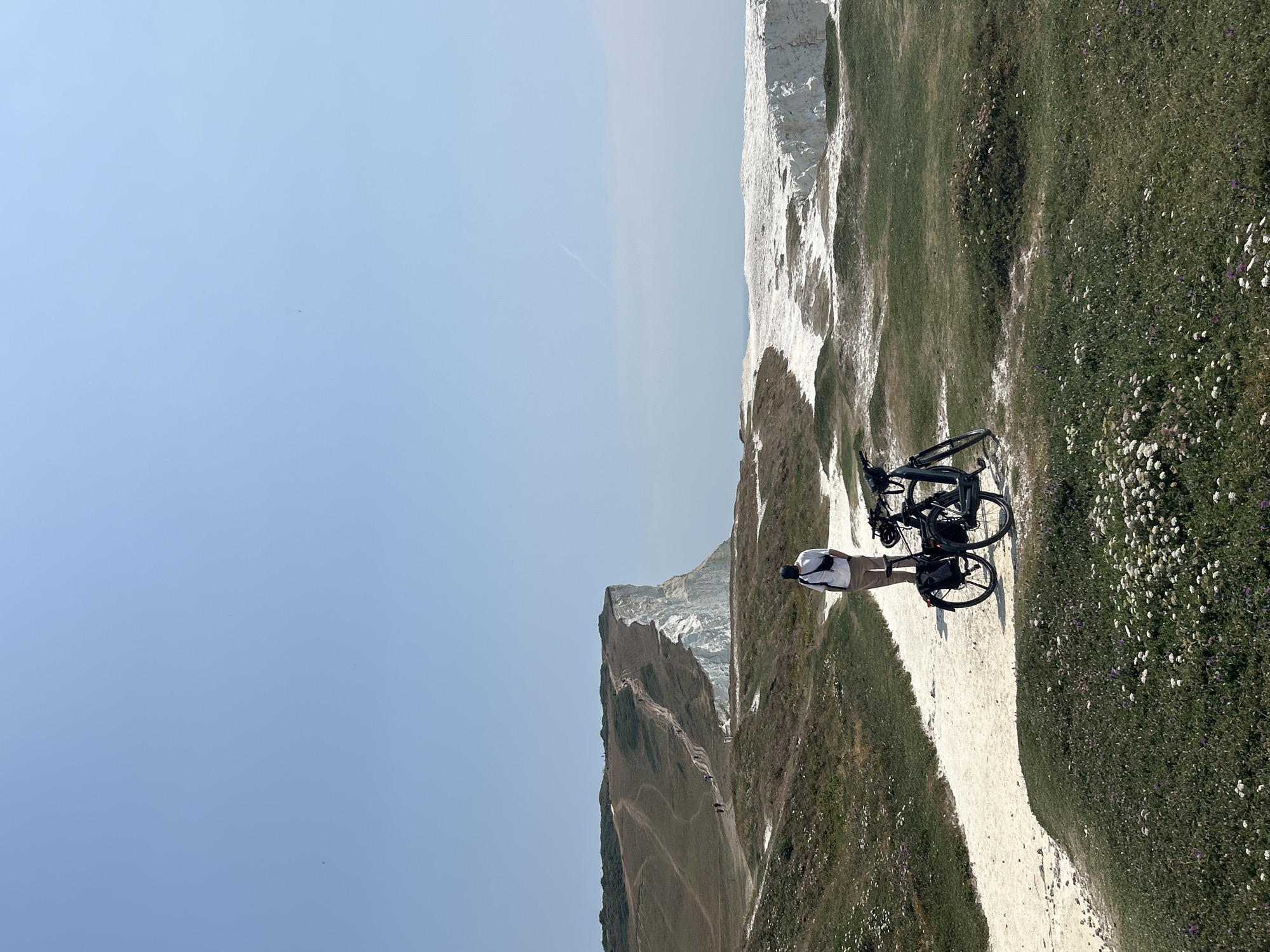 A person stands on a gravel path overlooking the Seven Sisters white cliffs in Seaford, United Kingdom. The individual, wearing a white t-shirt, beige trousers, and a dark cap, has a black cross-body bag. Two bicycles are parked on the path next to the person. The foreground is covered in green grass with scattered small white and purple flowers. The path leads towards the dramatic white cliffs, which are partially obscured by the terrain. Several other people can be seen walking along a path on the grassy hillside in the distance, leading up to the cliff edge. The sky is clear and blue.