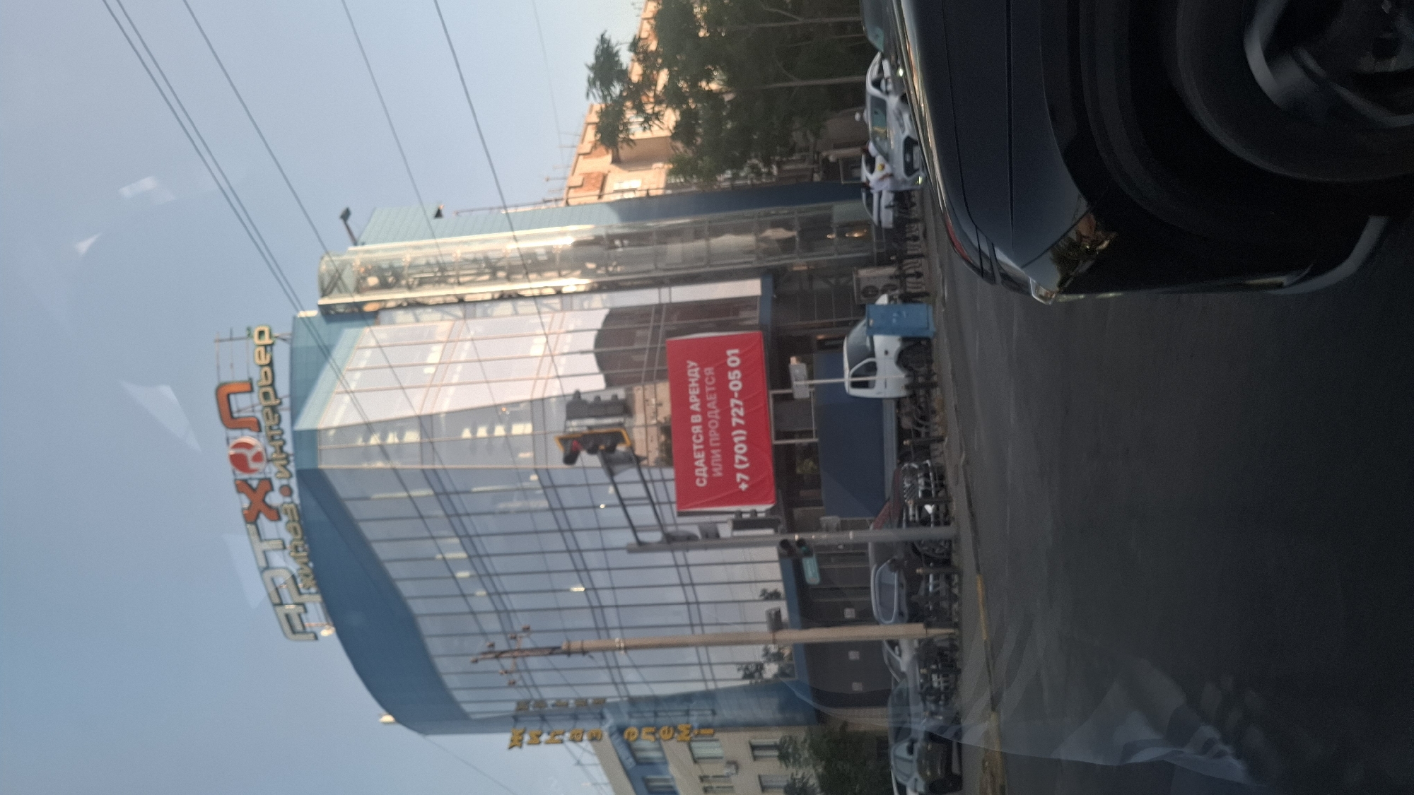 An exterior, eye-level, full shot captures a modern, glass-faced commercial building situated in Shymkent, Kazakhstan. The building's facade is dominated by blue and clear glass panels, reflecting the sky and surrounding environment. Atop the building, a prominent sign in red and orange letters reads "АРТХОЛ" with smaller text below it. A large red banner is affixed to the building, displaying a message in Russian that translates to "FOR RENT OR FOR SALE" and a phone number: "+7 (701) 727-05 01". A traffic light hangs near the banner. Several vehicles, including cars and a truck, are visible on the street in front of the building. The sky is clear and a pale blue, suggesting it is daytime. The scene is static, depicting a typical urban streetscape.