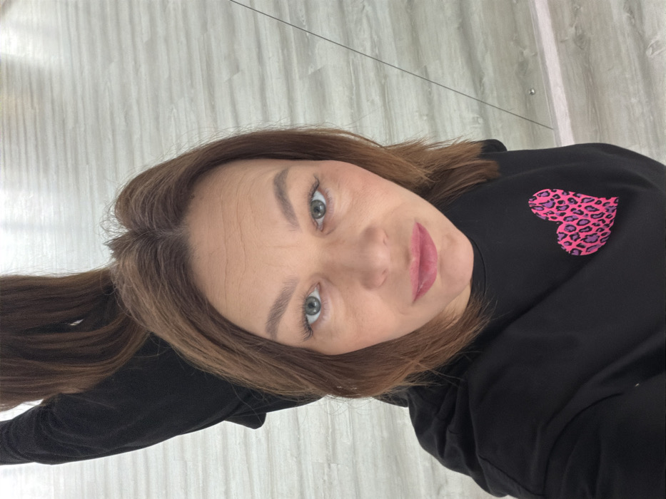 An adult female is depicted in a close-up, head-and-shoulders shot, appearing to be a selfie taken into a large reflective surface such as a mirror. She has medium-brown, shoulder-length hair parted in the middle, and light-colored eyes. Her face shows some forehead lines and she is wearing pink lipstick and eye makeup. She is dressed in a black long-sleeved top or sweatshirt featuring a prominent bright pink, leopard-print heart graphic on the left chest area. A blurred reflection of her back and hair is visible in the upper left of the image, indicating the reflective nature of the surface she is photographing herself in. The background reveals a light-colored floor, possibly wood-effect laminate or tile, with distinct horizontal lines. This scene is observed within the city of Chaikovsky, Russia.
