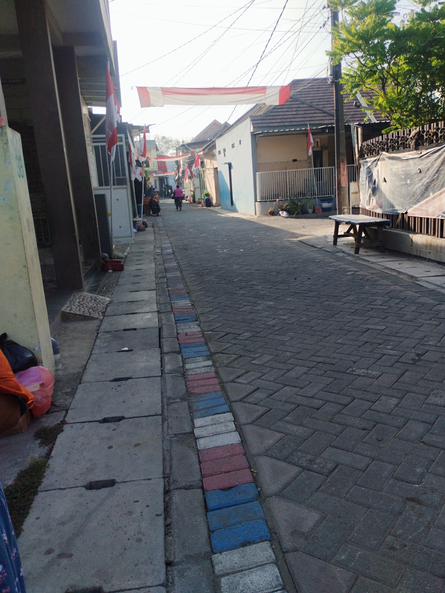 The image shows a street in the city of Surabaya, Indonesia, decorated with Indonesian flags and banners. The street is paved with interlocking bricks, and there are buildings on both sides. A few people are visible on the street, some on motorcycles and one on a bicycle. The sun is shining, casting shadows on the street. The scene suggests a festive occasion, possibly a national holiday.