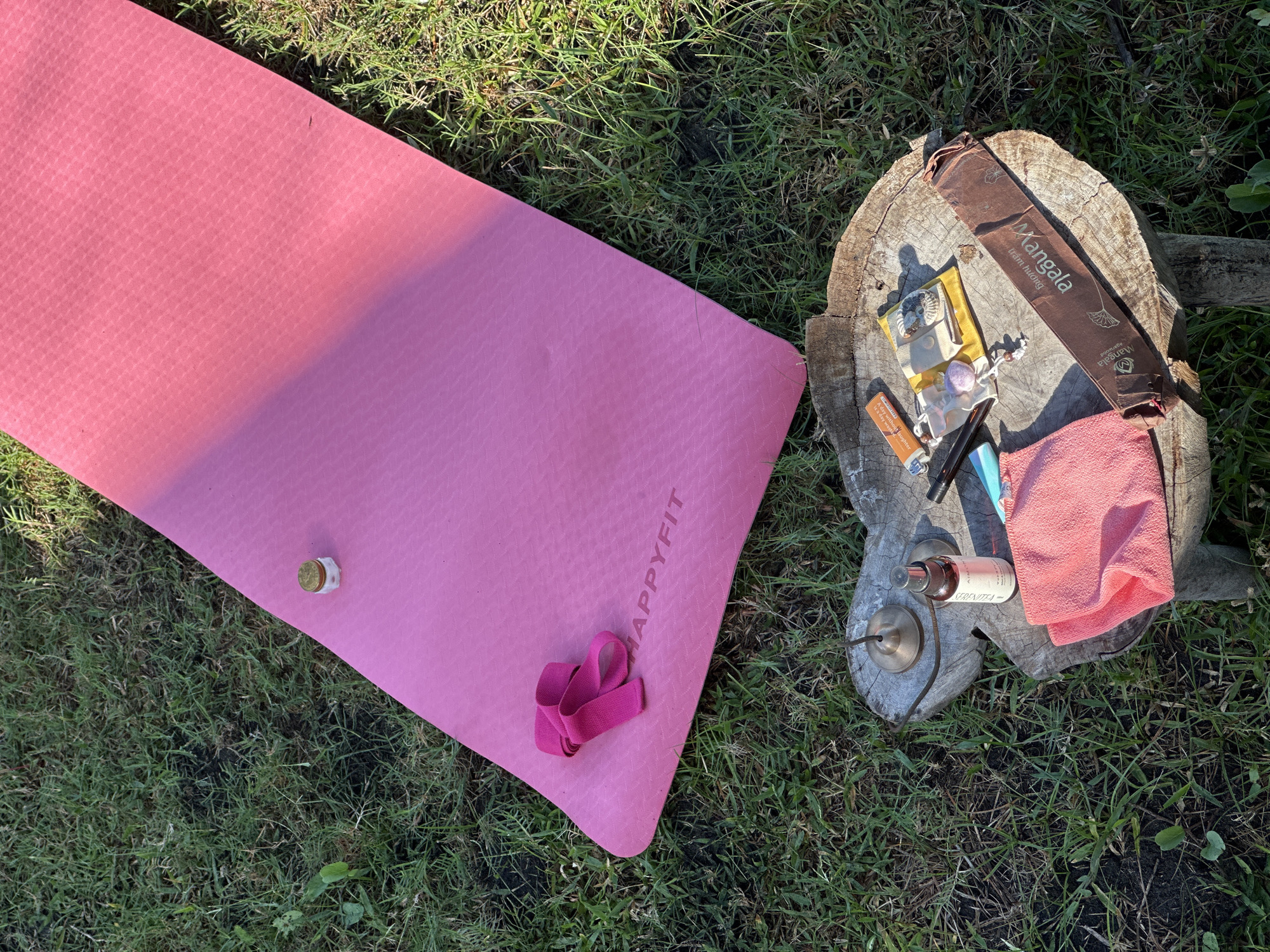 The image shows a pink "HappyFit" yoga mat on a grassy lawn. A small, lidded jar sits on the mat near the top. Next to the mat is a weathered tree stump on which various items are arranged: a brown box labeled "Xangala" with incense inside, a lighter, a small yellow bag, crystals, and a vial with a dropper. Also on the stump are a small towel, a spray bottle, and a small cymbal. The scene suggests an outdoor yoga and meditation setup. No people are visible. Mendoyo, Indonesia, is not identifiable as the specific location from the image itself.