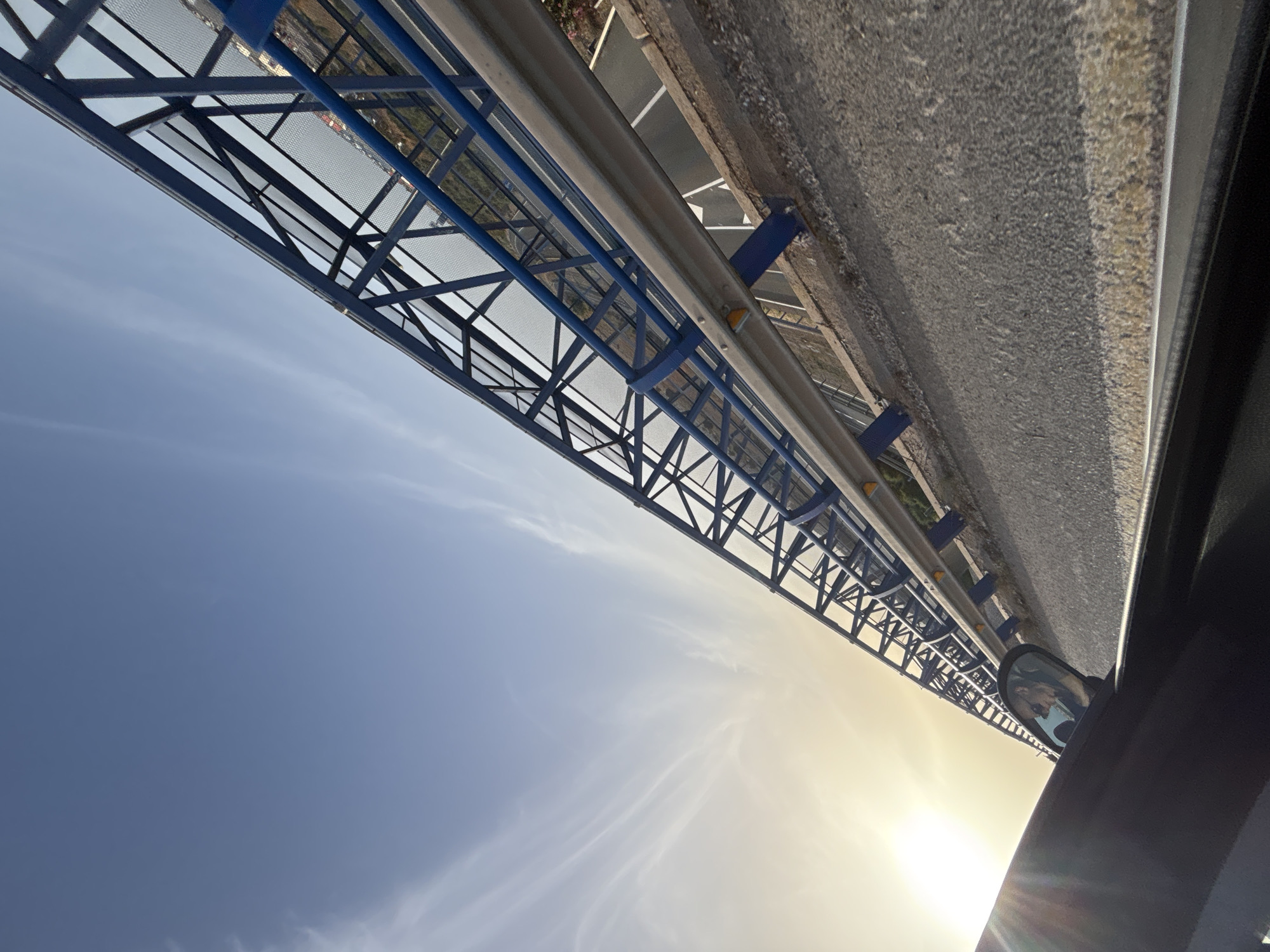 The image depicts a view from a car traveling on a road that appears to be elevated, likely a bridge or overpass. The sky is a clear blue with wispy white clouds and the sun is setting or rising on the left side of the frame, casting a warm glow. A prominent blue metal structure, possibly a sound barrier or part of a pedestrian bridge, runs parallel to the road, extending into the distance. The road itself is asphalt with white and yellow markings. In the side mirror of the car, a reflection of a person wearing sunglasses and a hat is visible. The overall scene suggests a journey on a highway or a similar elevated roadway in or near Orihuela, Spain, characterized by the distinctive blue barrier and the bright, possibly Mediterranean, sky.