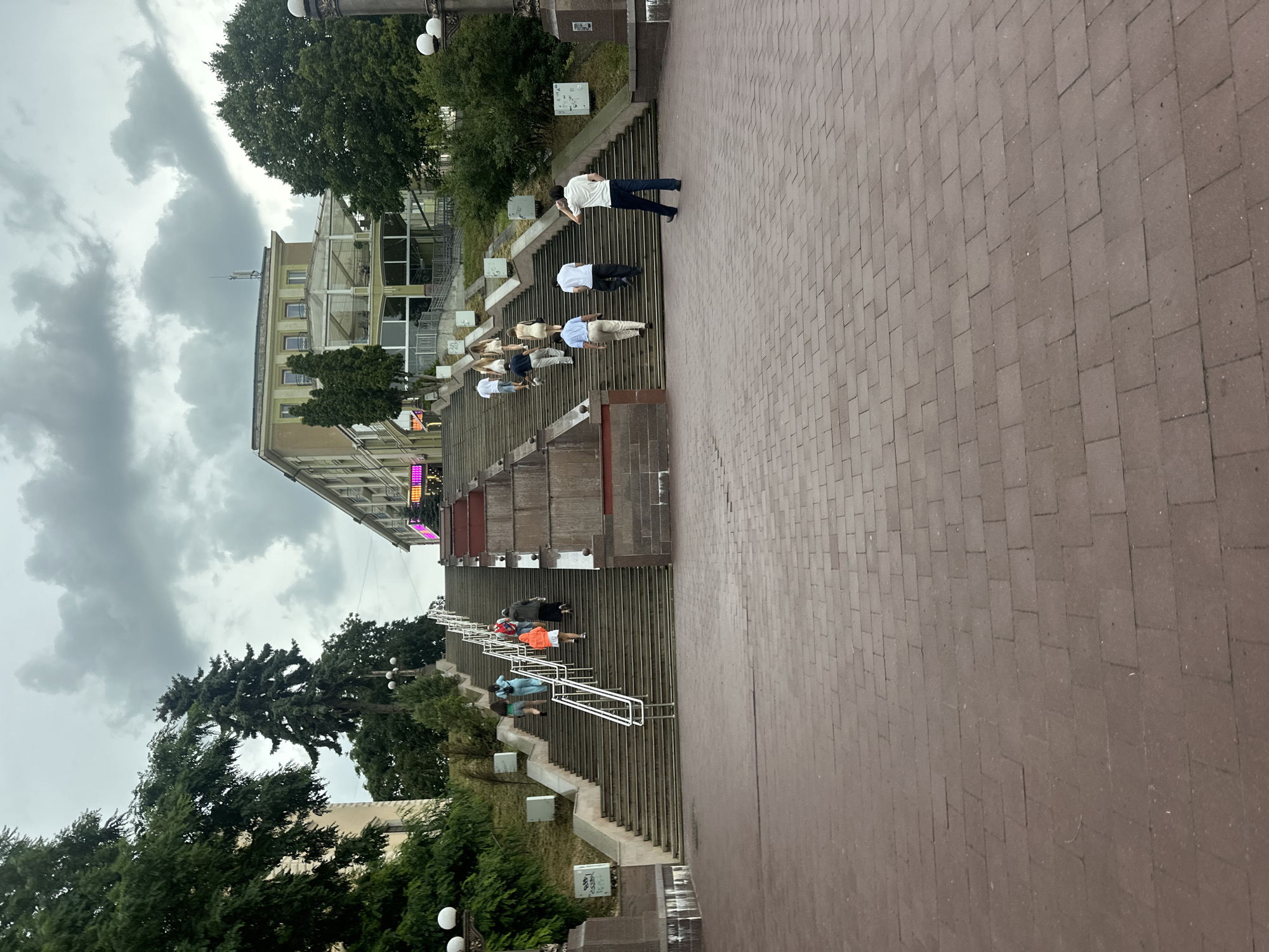 The image shows a wide, outdoor staircase in Ternopil, Ukraine, leading up to a building with a terrace. Several people are ascending the stairs. In the foreground, a man in a white polo shirt and dark pants walks away from the camera. To his left, a group of people are climbing the stairs, including a woman in an orange jacket. Further up, more individuals are visible, dressed in casual attire. The staircase is made of stone and has metal handrails. The surrounding environment includes trees and a cloudy sky. The building at the top appears to be a restaurant or cafe.