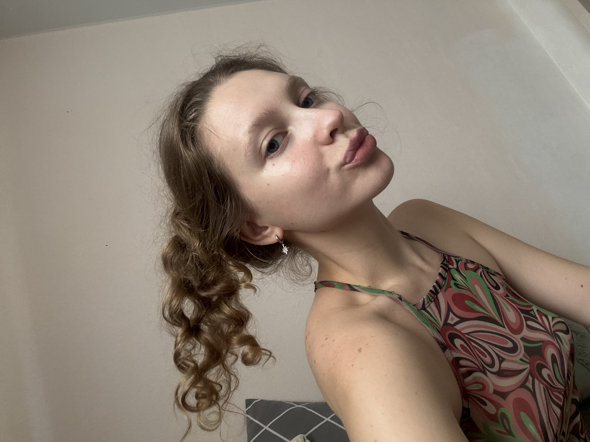 The image shows a young woman with light brown, curly hair pulled back into a ponytail. She is wearing a colorful, patterned top with thin straps. She is making a kissing face, with her lips pursed. A small, star-shaped earring is visible in her left ear. The background is a plain, light-colored wall. No specific location within Izhevsk, Russia, can be identified from the image.