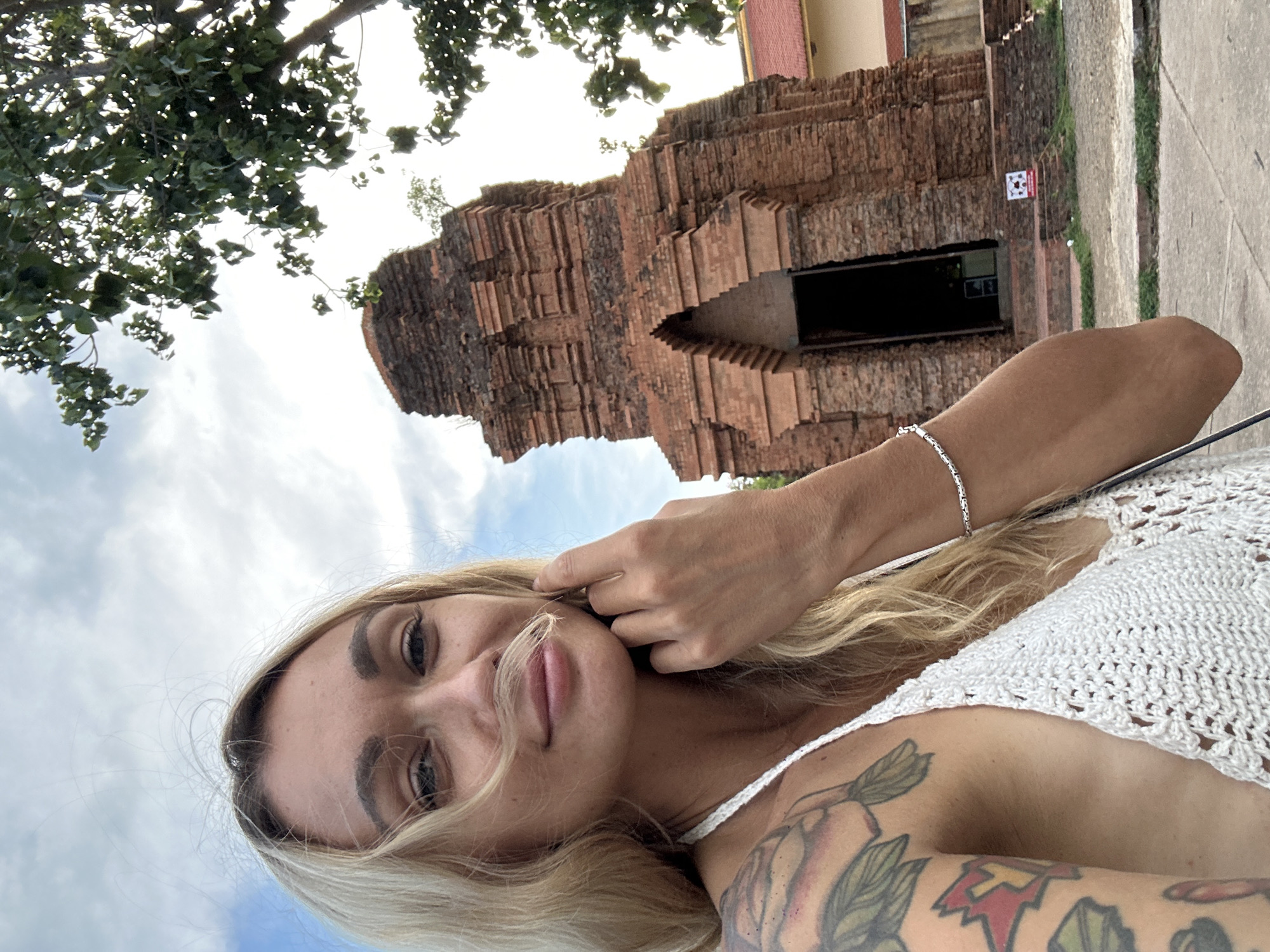 A fair-skinned person with long blonde hair, wearing a white crochet top and a silver bracelet, takes a selfie. The person has a shoulder tattoo with flowers and symbols. In the background is a brick structure, likely a temple or historical building. The photo appears to be taken outdoors, possibly in Phu Thuy Ward, Vietnam, based on potential location. Above the structure, there is a tree and a sky with clouds.
