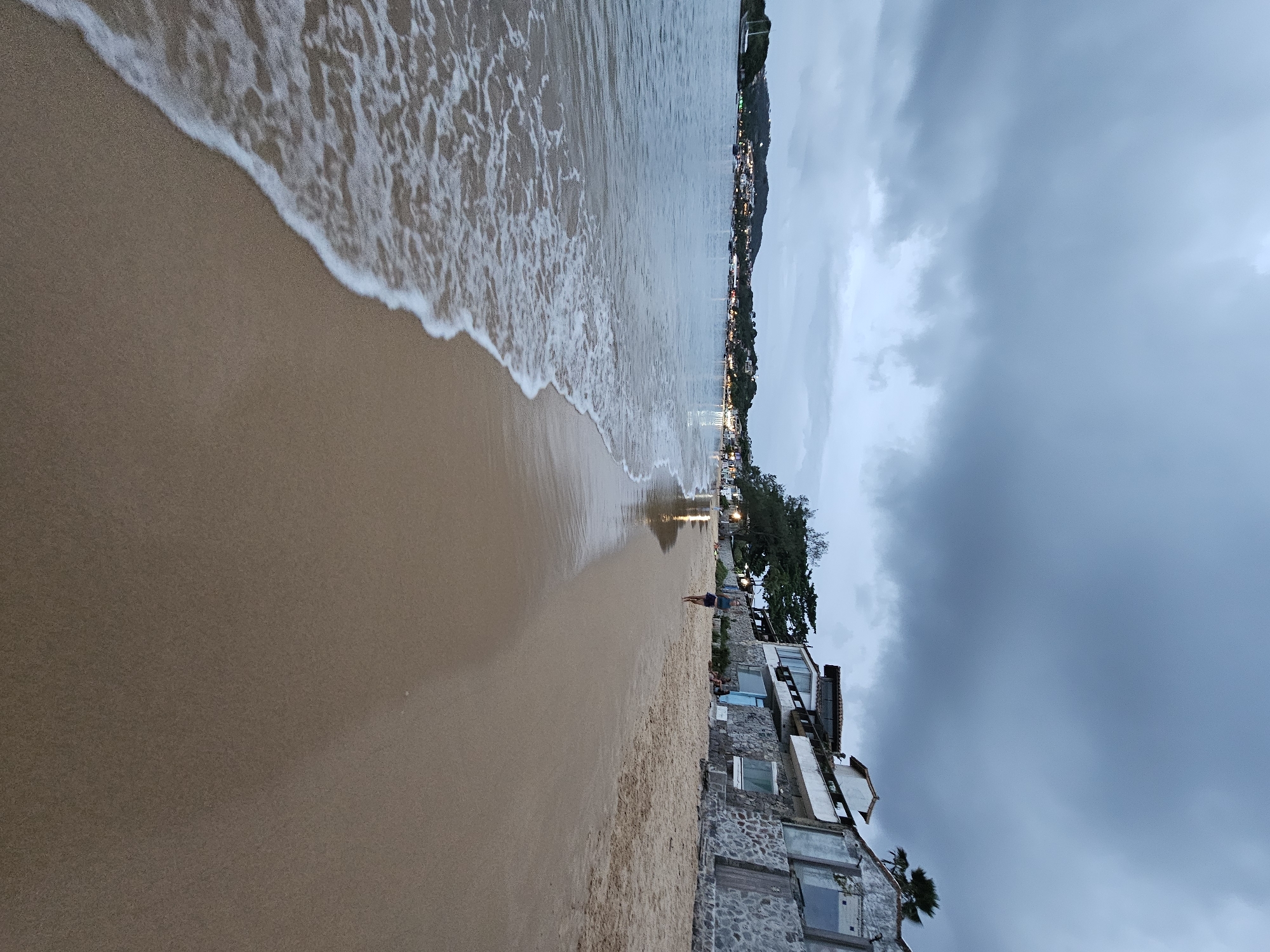 The image shows a beach in Armação Dos Búzios, Brazil, under a cloudy sky. The scene captures the shoreline with waves gently washing onto the sand. To the right of the frame, there's a building constructed with stone, situated close to the beach. A few people can be seen on the beach in the distance. The background features a line of lights suggesting urban development along the coast.