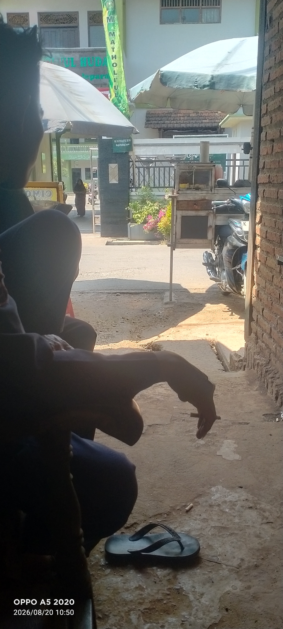 The image depicts an outdoor scene in Bugel, Indonesia, on August 20, 2026, at 10:50 AM. In the foreground, partially silhouetted figures are seated. One figure has their hand extended, holding a cigarette. Nearby, a pair of dark flip-flops rests on the ground. The ground is a mix of dirt and concrete, with some patches of lighter material.
In the middle ground, a street is visible, with a woman in dark clothing walking away from the camera. Adjacent to the street is a small food cart or stall, with a motorcycle parked behind it. The cart appears to be made of wood and glass, with some items displayed inside. There is also greenery, including flowering plants, on the right side of the street.
In the background, the facade of a building is visible, featuring Arabic calligraphy and architectural details. A green banner with text in Arabic and Indonesian, reading "MATHOLUL ULUM," is prominently displayed. The building appears to be an educational institution, possibly a school or Islamic boarding school. Umbrellas or awnings are also visible, providing shade. The overall lighting suggests a sunny day. The brick wall on the right side of the image frames the scene.