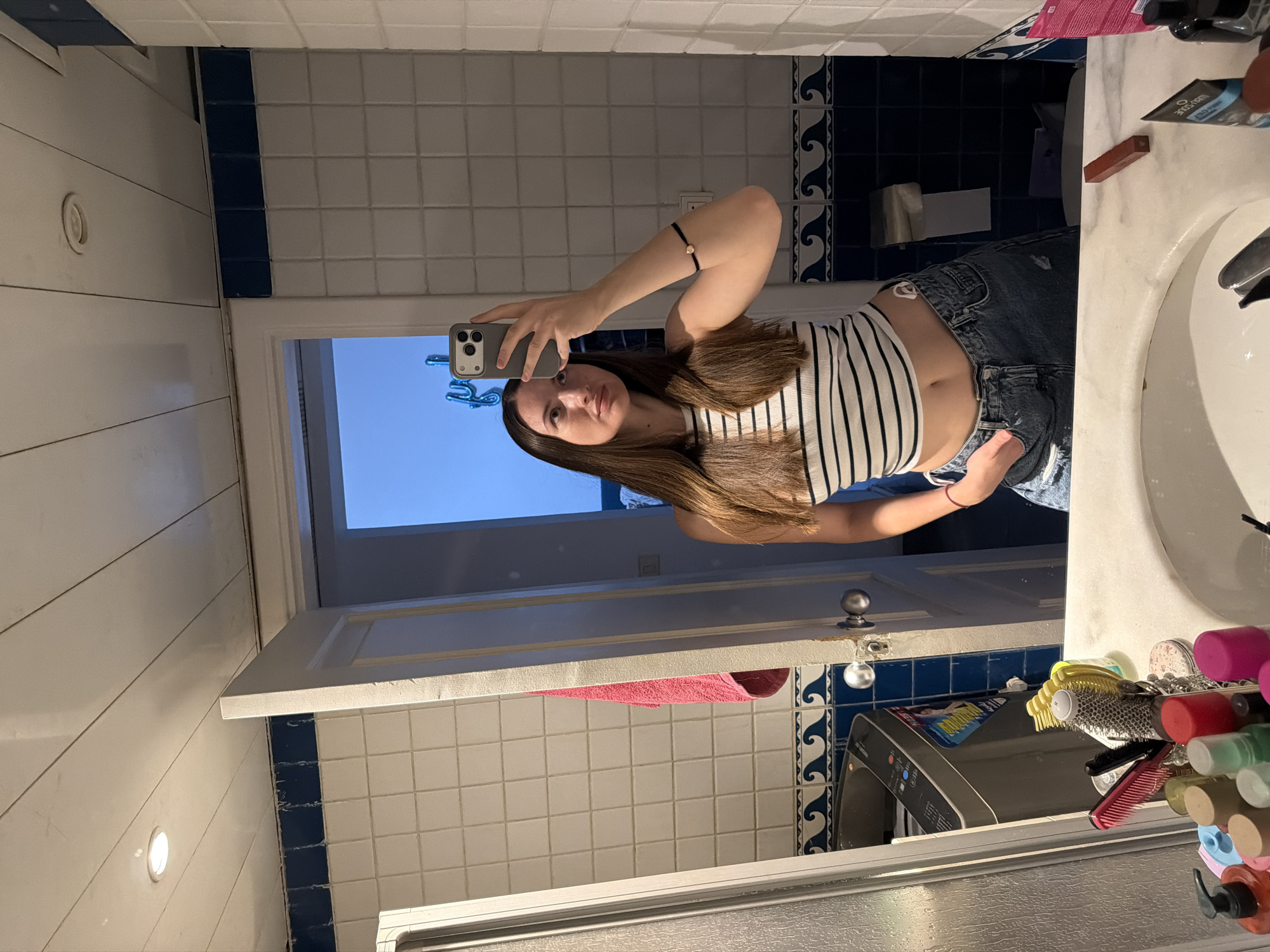 A young woman with long, brown hair is taking a mirror selfie in a bathroom. She is wearing a black and white striped crop top and distressed denim jeans. She is holding a smartphone in her right hand, which is raised to take the picture. The bathroom has white tiled walls with blue accents and a blue wave pattern border near the ceiling. A pink towel is hanging on a hook to the left of the mirror. In the reflection, a washing machine is visible to the left, and a partially open door reveals a glimpse of the sky outside. There are various toiletries and hair accessories on the white marble countertop in the foreground. The location is described as Sharm El Sheikh, Egypt.