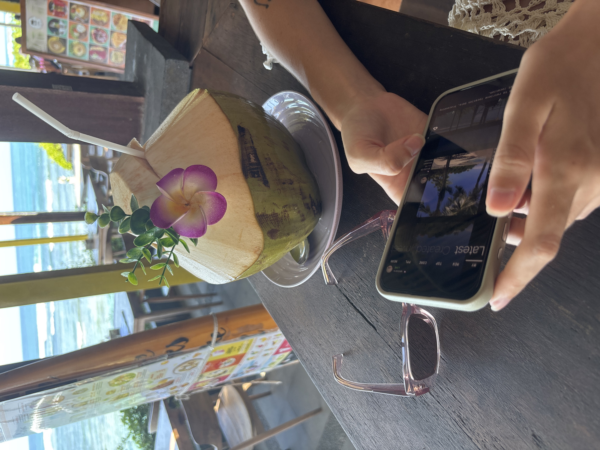 A person is holding a smartphone over a table with a coconut drink. The coconut drink is in a white plate. The drink has a straw and is decorated with a purple flower and green sprigs. A pair of glasses are on the table near the phone. The phone screen shows a picture of palm trees. The text on the screen says "Latest Created Invest." In the background, there is a view of the ocean and a building with a menu board. The location is likely Kuta Selatan, Indonesia, as the phone screen shows a photo from the beach of that city.