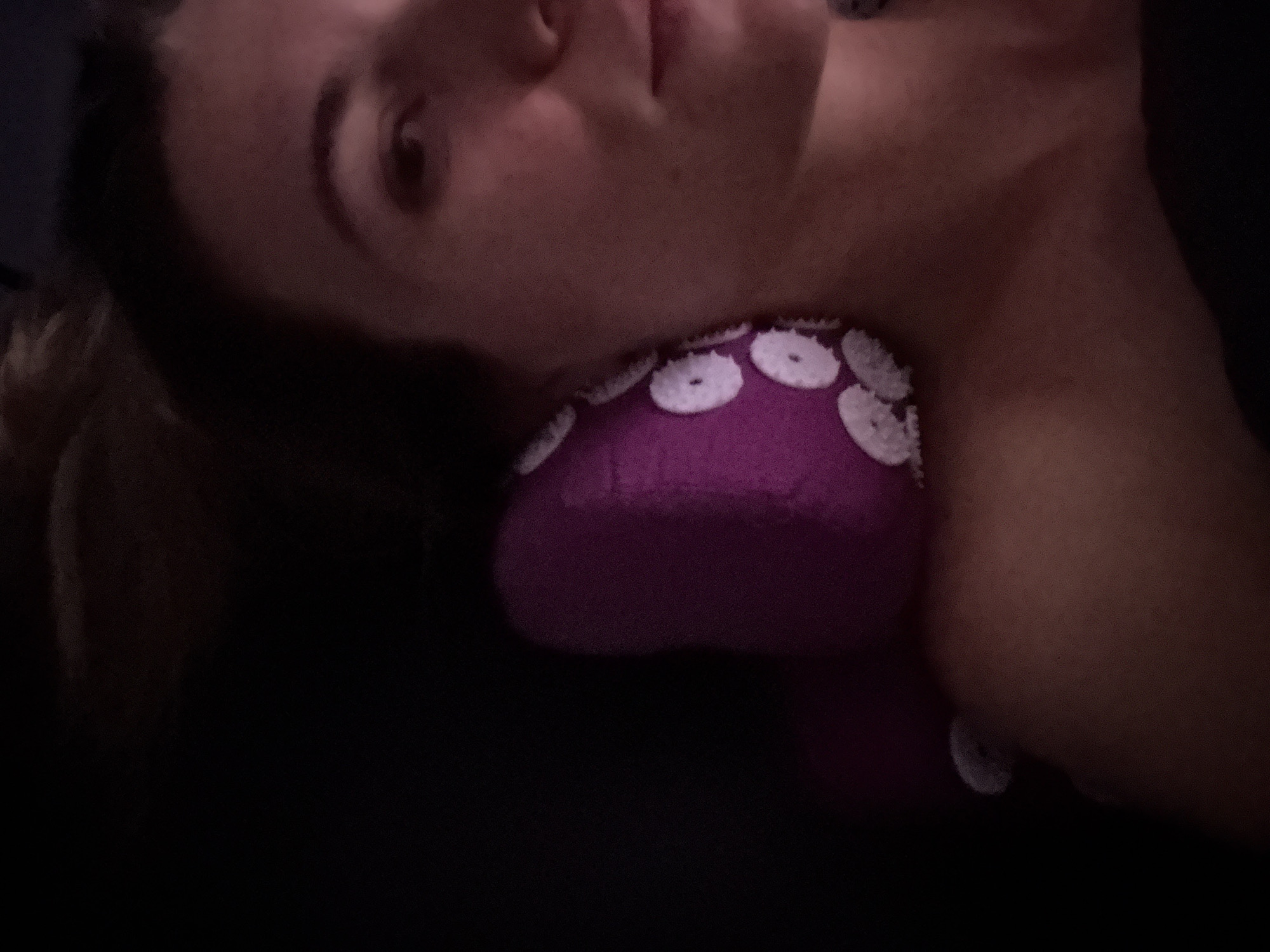 A person is lying down with their head resting on a purple, octopus-shaped pillow with white button-like details. The scene is dimly lit, making it difficult to discern specific details about the environment. The location is unknown.