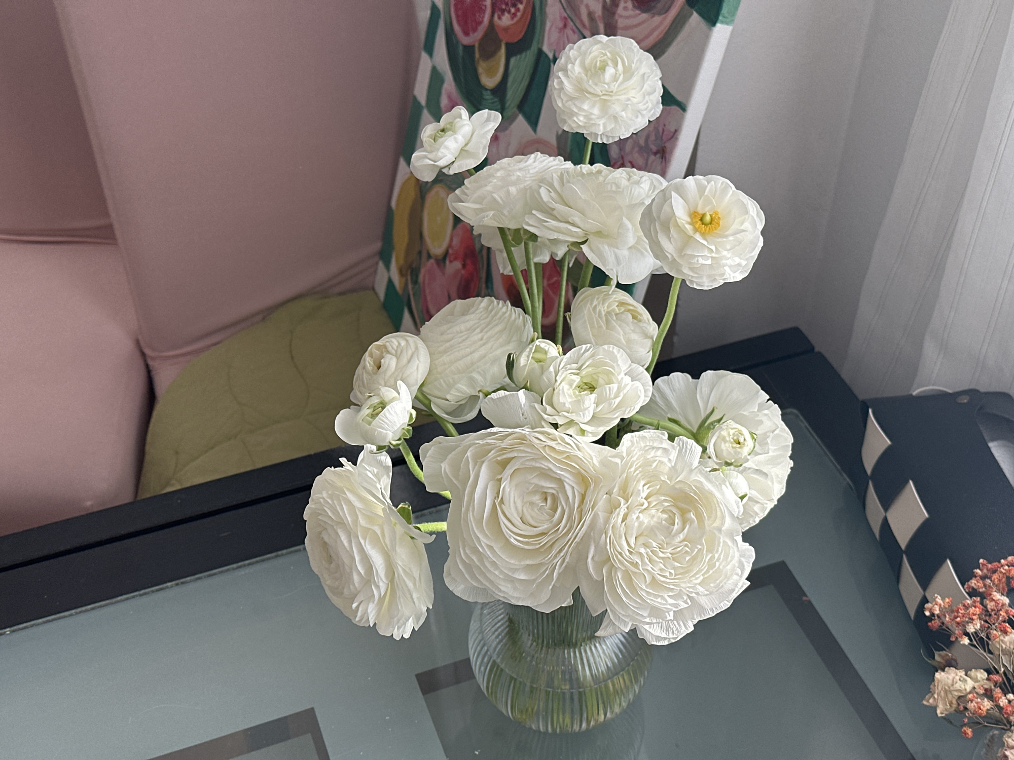 A close-up shot depicts a clear, ribbed glass vase filled with white ranunculus flowers arranged on a glass-topped table. The table, with a black border, sits near a pink upholstered sofa with a green cushion and a patterned piece of art leaning against the wall behind. A black object with white geometric patterns is visible on the right edge of the table near dried floral sprigs. The scene is well-lit, suggesting daylight. No people are visible in the frame. There is no way to determine the location is in the city of Novi Sad, Serbia.