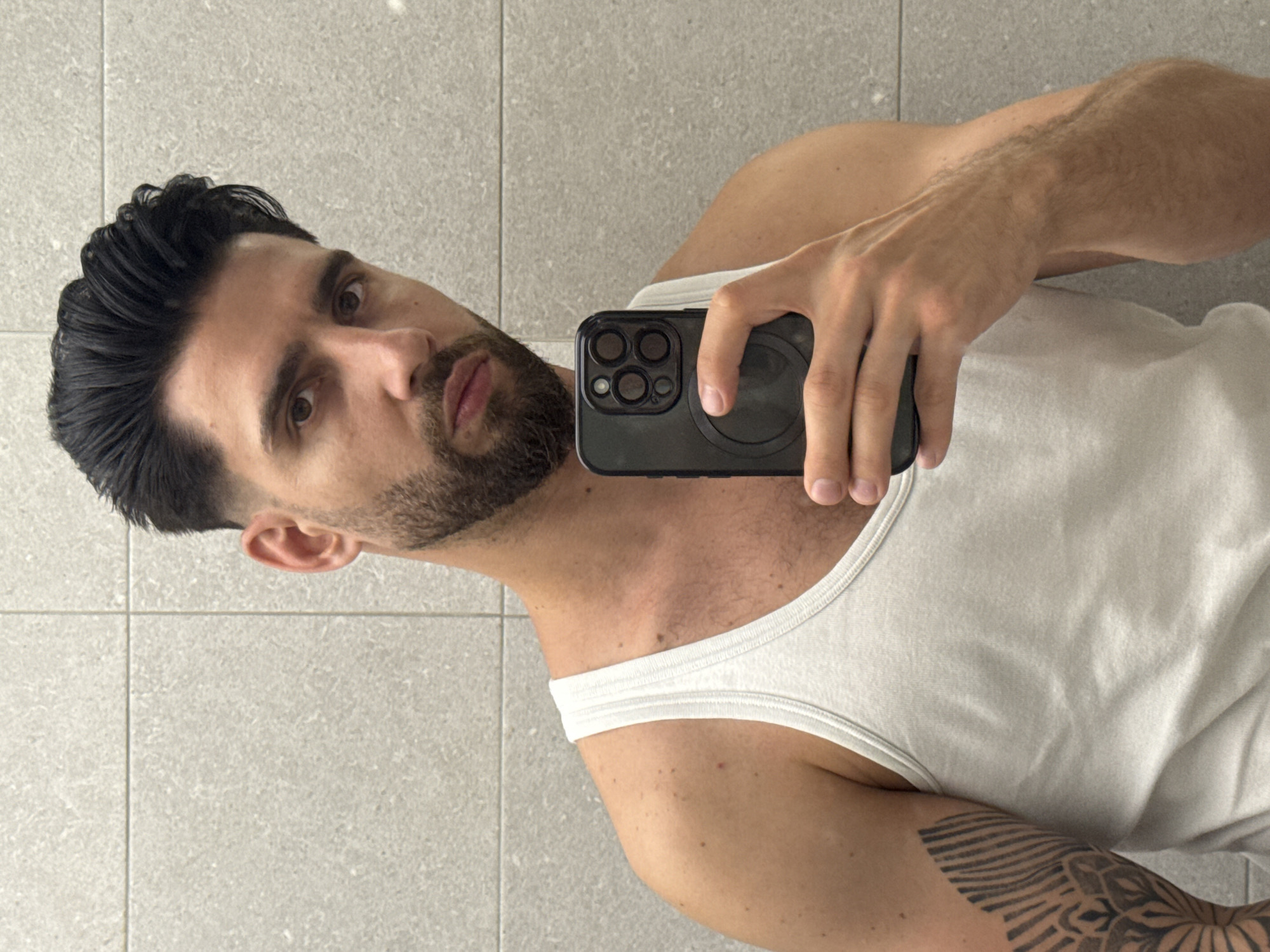 A man with dark hair and a beard is taking a selfie in front of a gray tiled wall, likely in a bathroom. He is wearing a white tank top and holding a dark-colored smartphone. He has a tattoo on his left arm. There is no information to suggest that this image was taken in Sofia, Bulgaria.