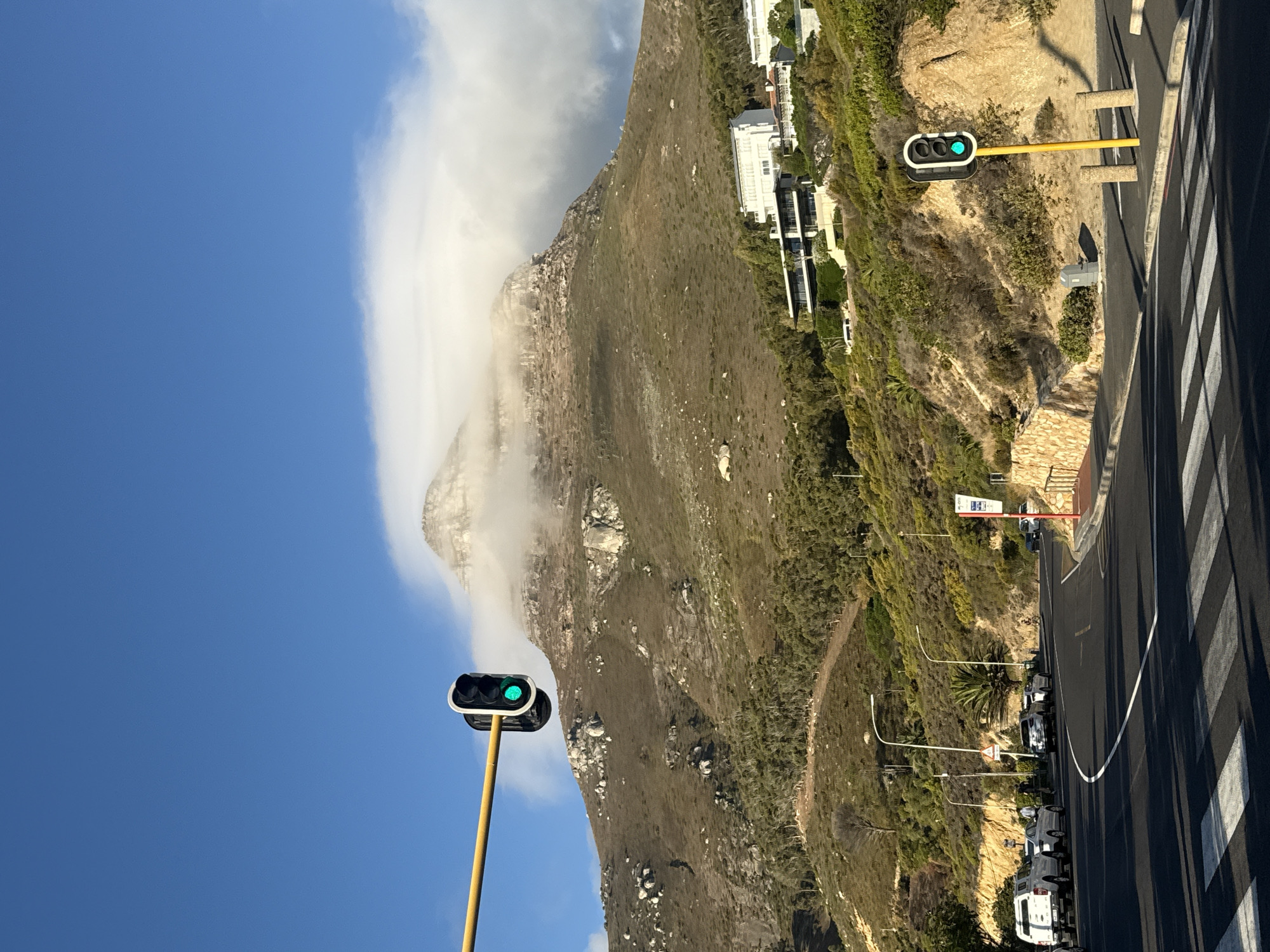The image displays a daytime scene in Cape Town, South Africa, featuring Lion's Head mountain dominating the background. The mountain's rugged, rocky summit and upper slopes are partially enveloped by a thick layer of white cumulus clouds, contrasting with the clear, deep blue sky above. The mid and lower slopes of the mountain are covered in sparse, dry olive-green and brown scrub vegetation, dotted with numerous grey boulders. Several modern, multi-level residential buildings with white facades and dark-framed windows are situated on the vegetated slopes, seamlessly integrated into the landscape.
In the foreground, a two-lane asphalt road, likely part of a coastal route such as Victoria Road near Camps Bay or Clifton, curves gently to the right. White pedestrian crosswalk stripes are painted across the lane closest to the viewer. Two traffic lights, supported by bright yellow poles, display green signals, indicating an open flow of traffic. One traffic light is mounted on a horizontal yellow arm extending from the left, positioned high above the road. The second traffic light stands vertically on the right side of the road.
On the left side of the road, several vehicles are stopped or parked, including a white pickup truck, a silver sedan, and a white SUV. Further down the curving road, additional vehicles are visible. Two individuals stand on the sidewalk near the parked vehicles on the left. A single person on a bicycle is observed further along the road on the right. The road is bordered by lush green and brown vegetation, including palm trees, and a textured stone retaining wall on the right embankment. Streetlights with curved poles and a red and white bus stop sign are also present along the road's edge.