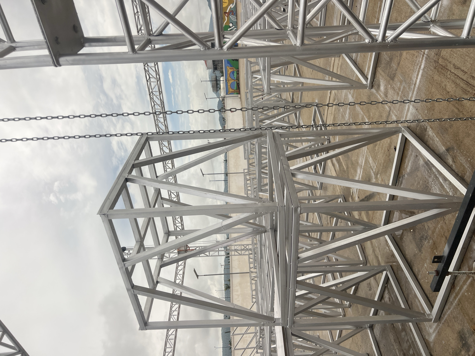 The image shows an outdoor scene with a large, complex structure made of interconnected aluminum trusses. The trusses are silver and form a grid-like pattern, creating a sense of depth and scale. There are several chains hanging vertically, attached to the trusses. In the background, there is a flat, paved surface, possibly a parking lot or an event ground, with some painted markings. Further in the distance, a low wall with some colorful graffiti is visible, and beyond that, there are trees and some hills under a cloudy sky. A few streetlights are also scattered across the scene. The overall impression is of a construction site or an area being prepared for an event, likely outdoors. The lighting suggests it could be daytime, with overcast or cloudy weather. No people are visible in the image, and there is no visible text. The location is identified as Howayi, Nigeria.
