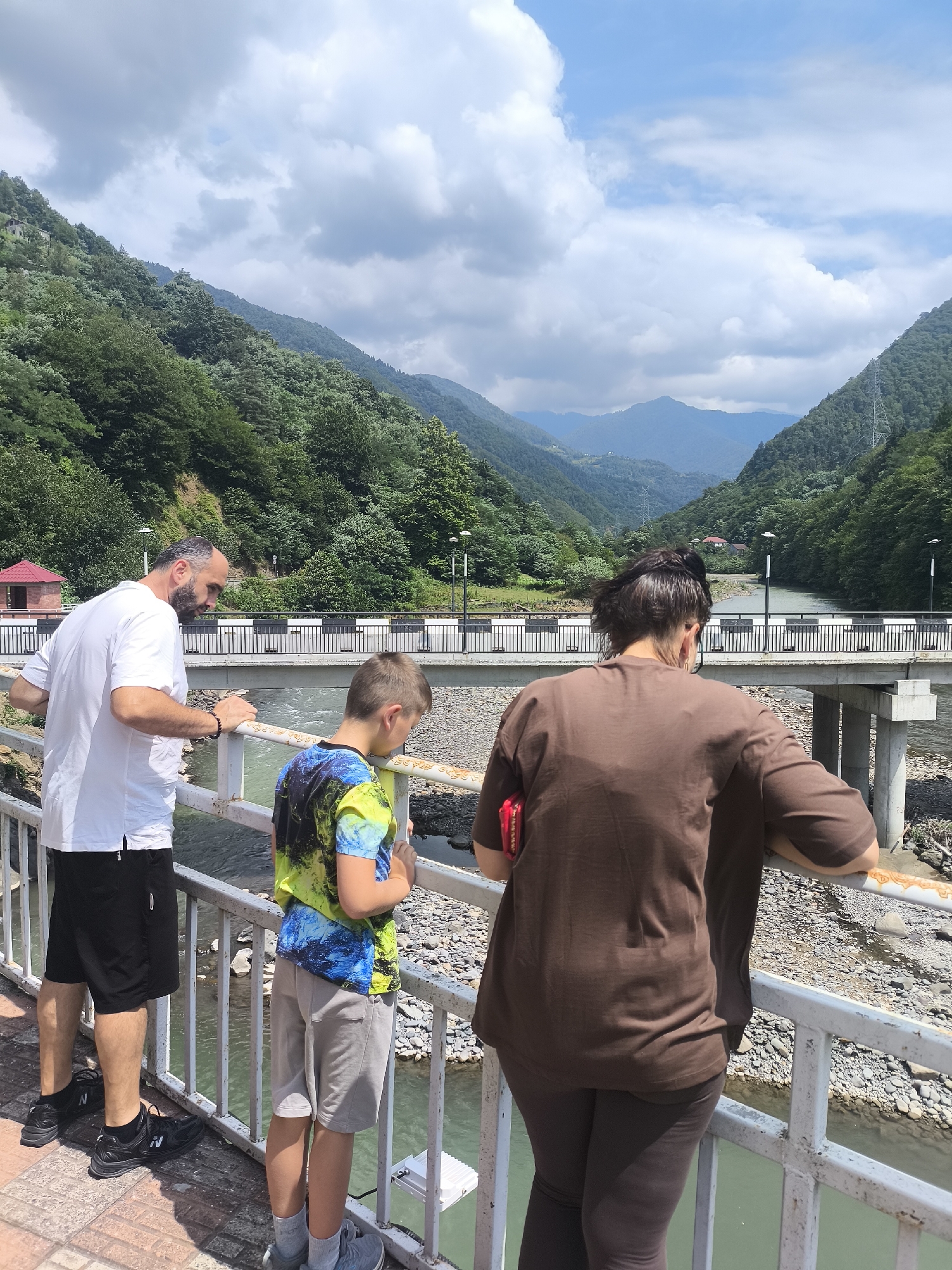 This is a vertical, eye-level, outdoor, full shot image taken on a cloudy day in Zvare, Georgia. Three people are standing on a white metal railing, looking out at a wide river valley.
On the left, a bald man with a dark beard, wearing a white t-shirt and black shorts, leans on the railing. Next to him, a young boy wearing a colorful t-shirt and light gray shorts looks down. To the right, a woman with dark hair pulled back, wearing a brown t-shirt and dark leggings, leans on the railing with her arms crossed.
The railing is white with a distressed appearance and some orange discoloration. The people are standing on a paved walkway with a red and brown brick pattern.
Beyond the railing, a wide, greenish river flows through a rocky bed. A concrete bridge with multiple pillars spans the river. The bridge has a white railing with black vertical bars. On the far side of the river, a small red-roofed building is visible.
The valley is steep and covered in dense green trees. In the background, more layers of mountains are visible under a sky with scattered white clouds.