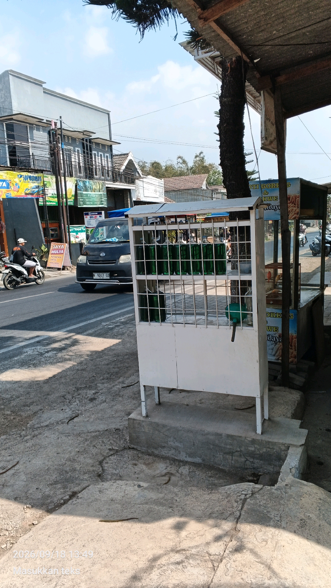 In Jeru, Indonesia, a street scene is depicted on a bright, sunny day. A roadside stall displays several green bottles of liquid, possibly fuel, within a white metal rack. To the left, a black van with license plate N 9247 GD is driving on the road, and a person on a white motorcycle is approaching. The background features buildings and trees, suggesting a commercial or residential area. The ground is a mix of concrete and asphalt, with some areas showing cracks and shadows from overhead structures. A timestamp at the bottom reads "2026/09/18 13:49".