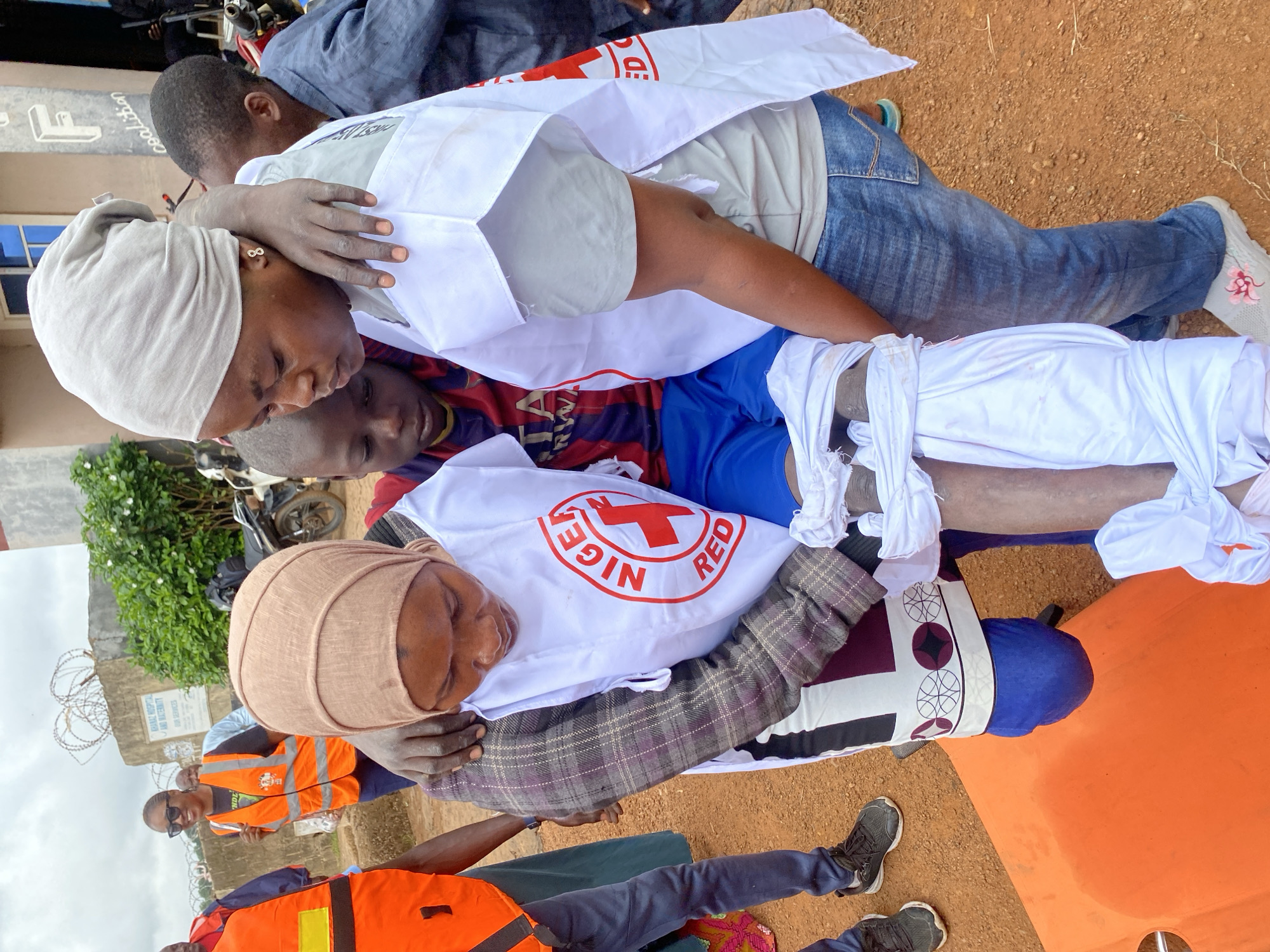 In Kawo, Nigeria, two women, identified by their Nigerian Red Cross vests, are assisting a young boy who appears to be injured. One woman, wearing a light grey headscarf and t-shirt, supports the boy's upper body. The other woman, in a brown headscarf and a plaid jacket over a white Red Cross vest, is also holding the boy. The boy is dressed in a red and blue striped shirt and blue shorts, with his legs wrapped in white cloth. They are positioned near an orange stretcher, suggesting they are preparing to move him. In the background, a sign on a building reads "KAWO HOSPITAL AND MATERNITY" and "OUR SERVICES." Other individuals and a motorcycle are also visible in the background. The ground is covered in reddish-brown dirt, characteristic of the environment in this region. The overall scene depicts a rescue or medical assistance operation.