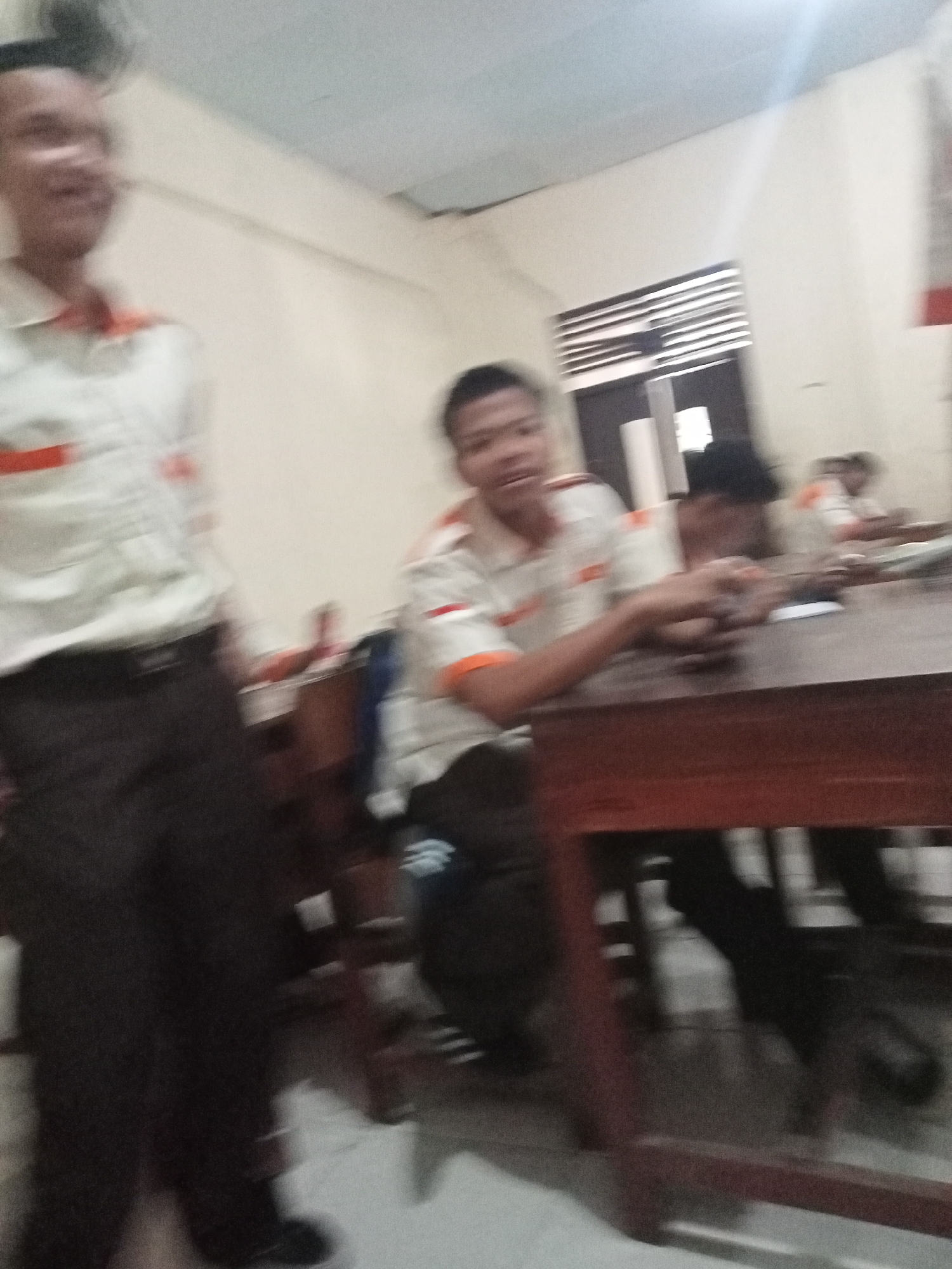 The image depicts a group of young males, likely students, in an indoor setting that appears to be a classroom or community hall in Secang, Indonesia. The environment is characterized by beige walls and a light blue ceiling, with a window featuring horizontal blinds visible in the background. A long wooden table occupies the foreground, with several individuals seated around it.
One individual in the foreground, on the left, is standing and appears to be wearing a light-colored shirt with orange accents and dark trousers. Another young male is seated at the table, facing the camera, wearing a similar shirt with orange detailing. His hands are resting on the table, and he seems to be holding or interacting with something small. Further back, other individuals are partially visible, also seated at tables, suggesting a communal or educational activity. The lighting suggests an indoor space, possibly illuminated by overhead lights or natural light filtering in from the window. The overall impression is one of a casual gathering or learning environment.