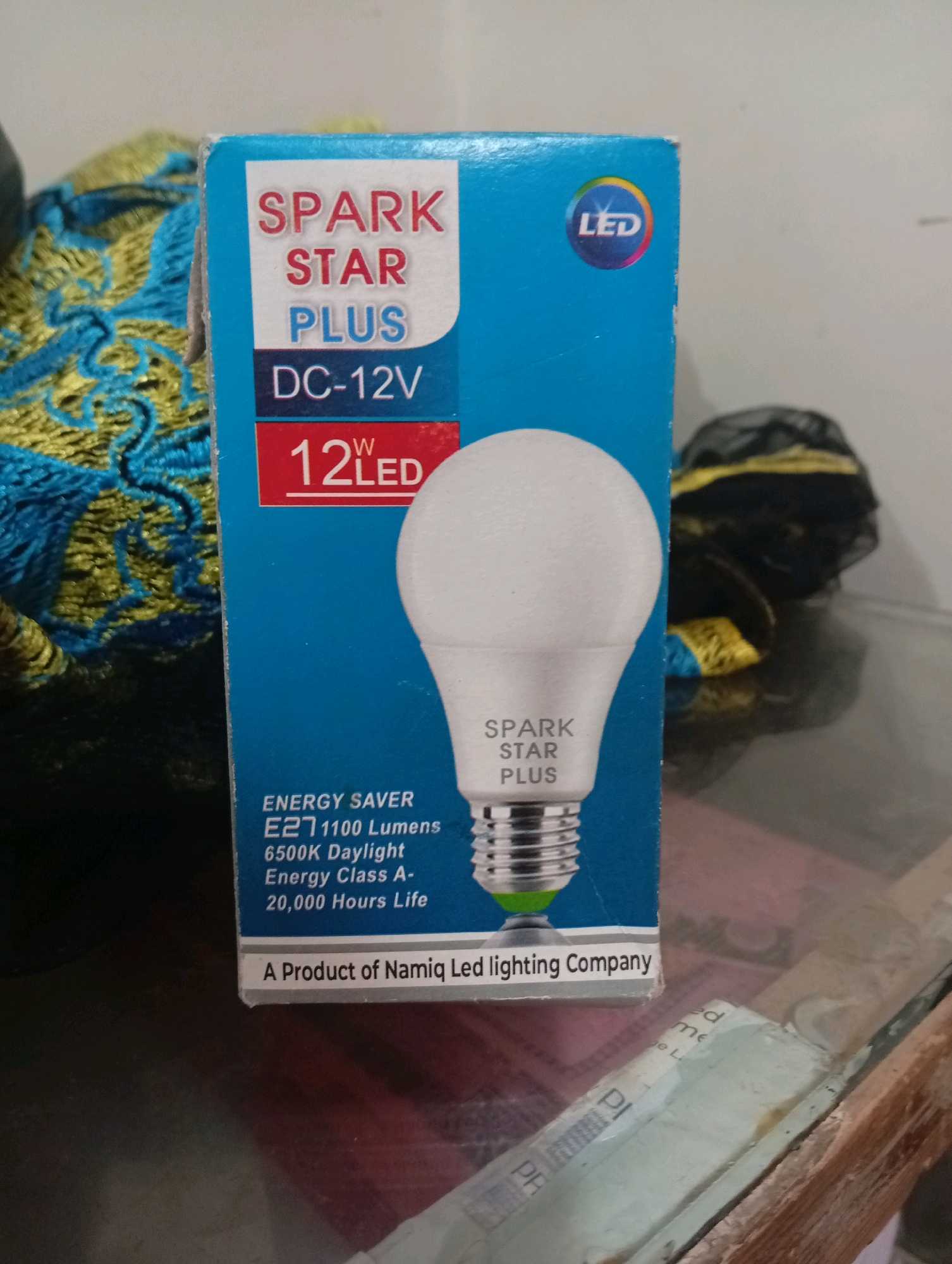 A product image shows a light bulb box with the brand "SPARK STAR PLUS" and "DC-12V" printed on it. The box also indicates "12W LED," "ENERGY SAVER," "1100 Lumens," "6500K Daylight," "Energy Class A-," and "20,000 Hours Life." At the bottom, it states "A Product of Namiq Led lighting Company." The box is partially in shadow. The background consists of a blurry textile with blue and gold patterns and a dark object. This image does not contain information about the city of Larkana, Pakistan.