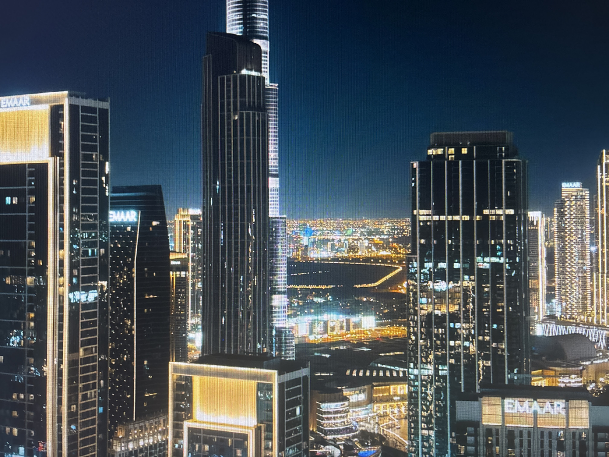 The image depicts a nighttime cityscape dominated by several tall skyscrapers. The most prominent structure is a very tall, thin tower located near the center of the frame. Many other illuminated buildings fill the midground, forming a dense urban environment. In the background, a network of bright lights stretches across the horizon. Each building is marked with "EMAAR" logo, signifying the name of the business. This is not the city of Durmersheim, Germany.
