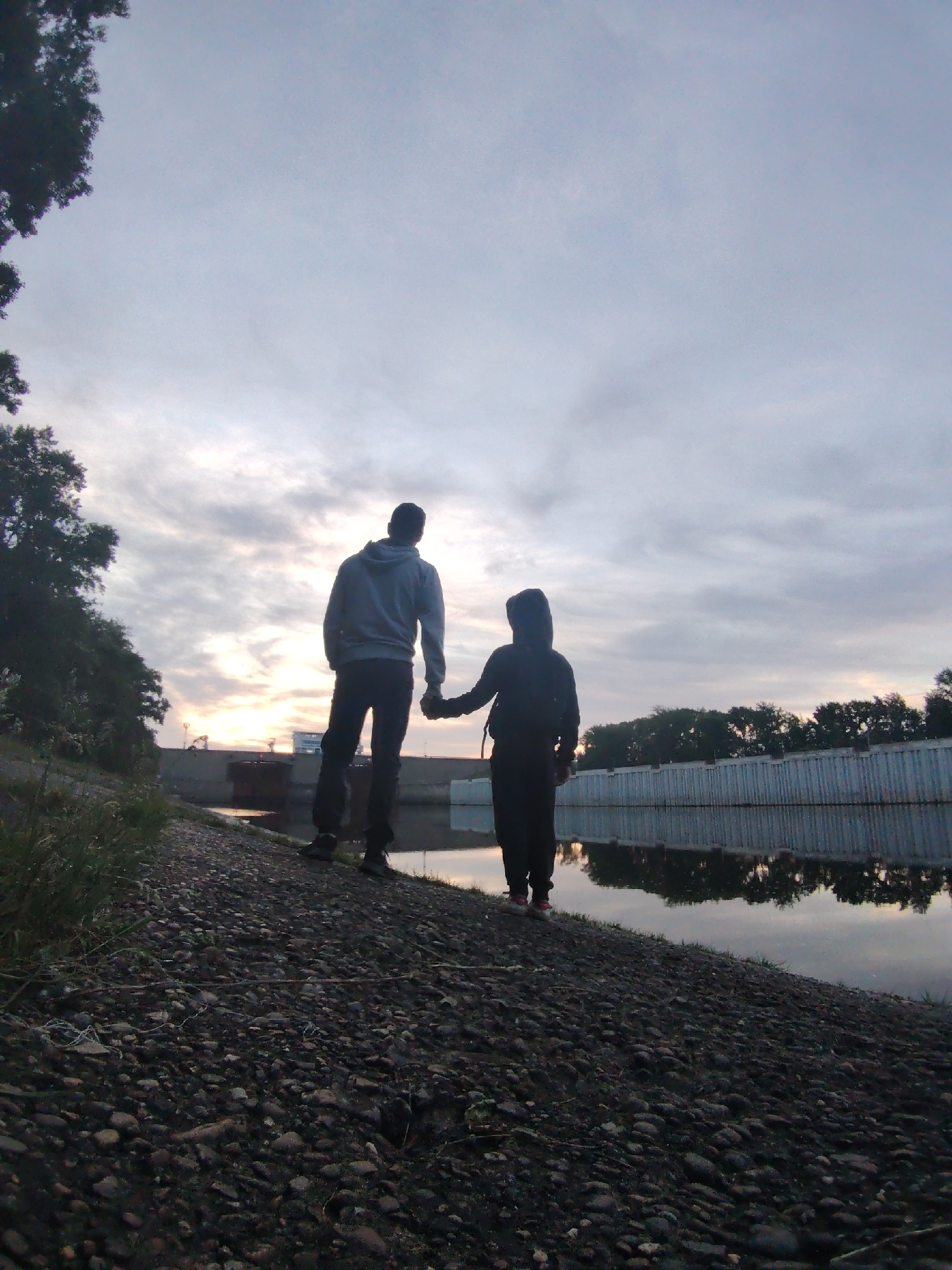 This image depicts two individuals, silhouetted and seen from behind, standing on the bank of a body of water, likely a river or canal, in Chaikovsky, Russia. The person on the left, appearing taller, wears a light-colored hooded sweatshirt and dark pants, while the shorter person on the right wears a dark hooded sweatshirt and dark pants. They are holding hands and gazing outwards towards the horizon.
The setting appears to be a riverbank with a dark, pebbled or gravelly ground in the foreground and beneath their feet, gently sloping down towards the water. Sparse patches of green grass are visible among the pebbles on the left. The water is calm and still, reflecting the soft, muted tones of the sky and distant trees.
The sky is largely overcast with soft grey clouds, but a faint, warm glow is visible low on the horizon between the two figures, indicating either sunrise or sunset. This atmospheric lighting contributes to a serene and contemplative mood. Dark foliage partially frames the upper left corner of the image. Across the water, a long, low retaining wall or barrier runs along the bank, with a line of trees silhouetted behind it. In the far distance, structures resembling parts of a bridge or industrial equipment are faintly visible against the slightly brighter sky.
The activity taking place is a shared moment of observation or reflection, emphasizing companionship through the gesture of holding hands. The scene evokes feelings of peace, connection, and appreciation for the tranquil, natural environment bordering an urban or industrial landscape. There is no visible text in the media.