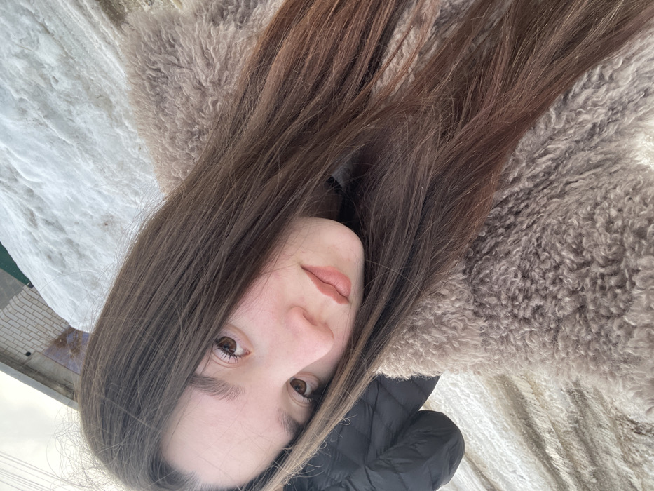 A young female subject is depicted in a close-up, tilted portrait outdoors in the city of Ольховка, Russia. Her long, straight, dark brown hair cascades down her front and shoulders. She has brown eyes, a fair complexion, and her lips are painted with a light, neutral-toned lipstick. She is wearing a light brown, shaggy-textured, possibly faux fur coat. A portion of a black quilted puffer jacket is visible on the lower left of the frame. In the background, a light-colored brick wall structure is partially visible to the upper left. A large accumulation of white snow with visible texture covers the ground behind and to the right of the subject, indicating a winter environment. The ground beneath the snow appears to be a mix of soil and debris. The sky is bright and overcast. The subject is looking directly at the camera.