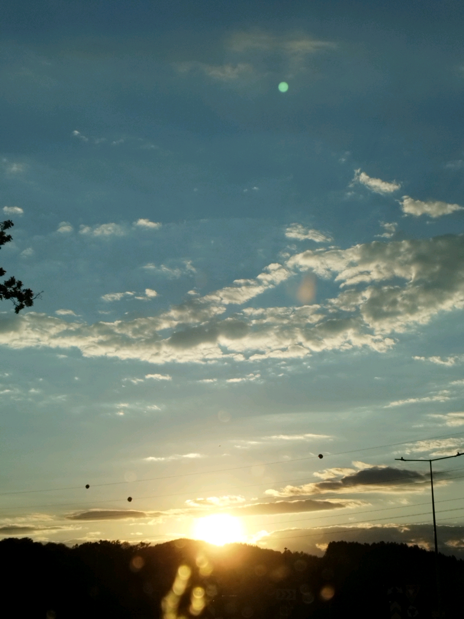 The image shows a sunset over a silhouetted landscape of hills and trees. The sky is a mixture of blue and gray, with scattered clouds illuminated by the setting sun. The sun itself is a bright, golden orb near the horizon, casting a warm glow across the scene. There are power lines stretching across the frame, with what appear to be two spherical objects hanging from them. A tall lamppost stands on the right side of the image, also silhouetted against the sky. The foreground is dark and out of focus, with some circular blurs that suggest it might be taken through a window. Based on the specific landmarks visible, such as the particular style of road signs and the surrounding topography, this scene is identifiable as being in the vicinity of Curtea de Argeș, Romania.