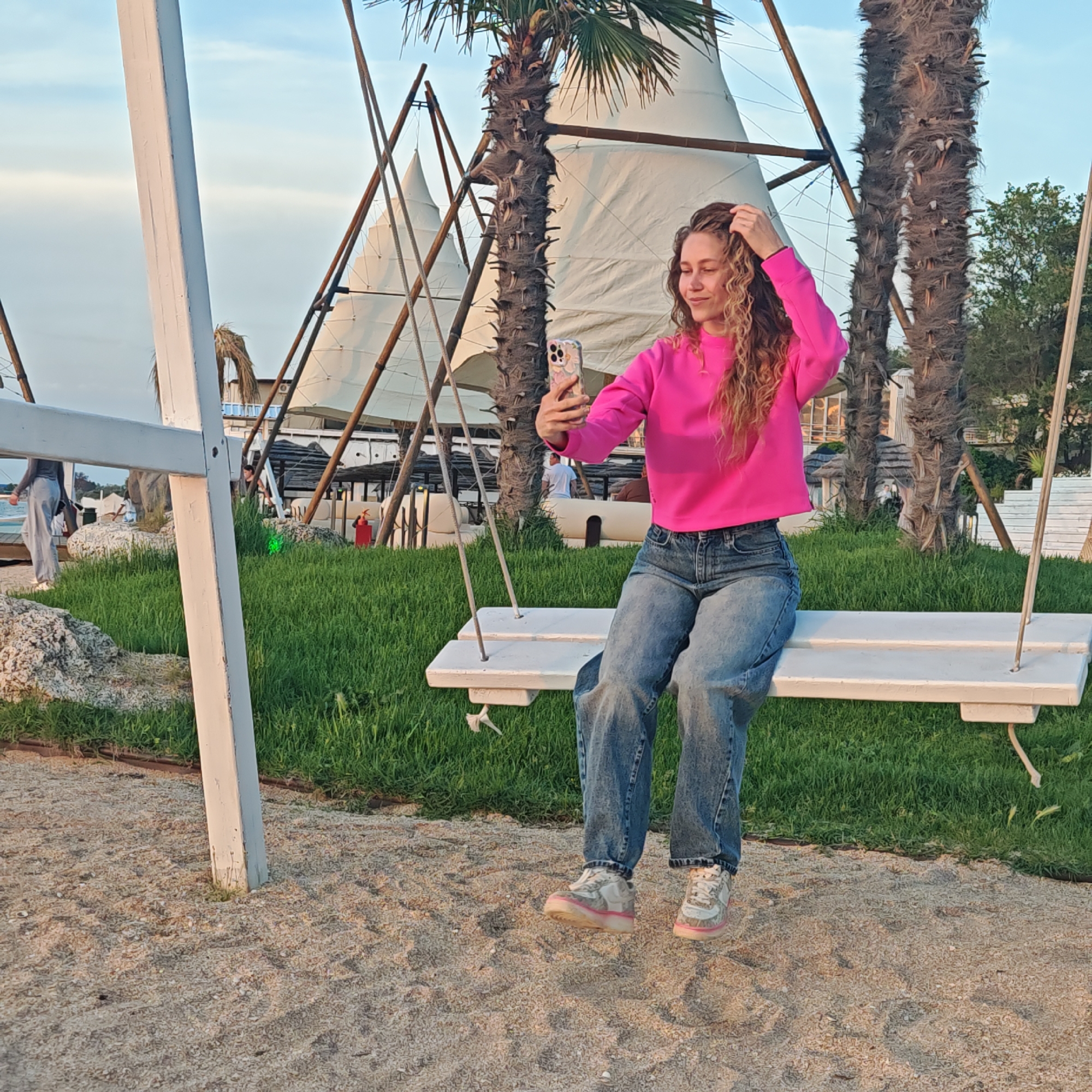 A woman with curly blonde hair sits on a white swing set at an outdoor location in Unknown, Unknown. She wears a bright pink long-sleeved shirt and blue jeans. She is holding a cell phone in her right hand, apparently taking a selfie, and her left hand is in her hair. The swing set is on a sandy surface, with green grass growing nearby. Behind the swing set are tall, cone-shaped structures made of white fabric supported by wooden poles. There are also tall palm trees in the background. The sky is visible in the upper portion of the image.