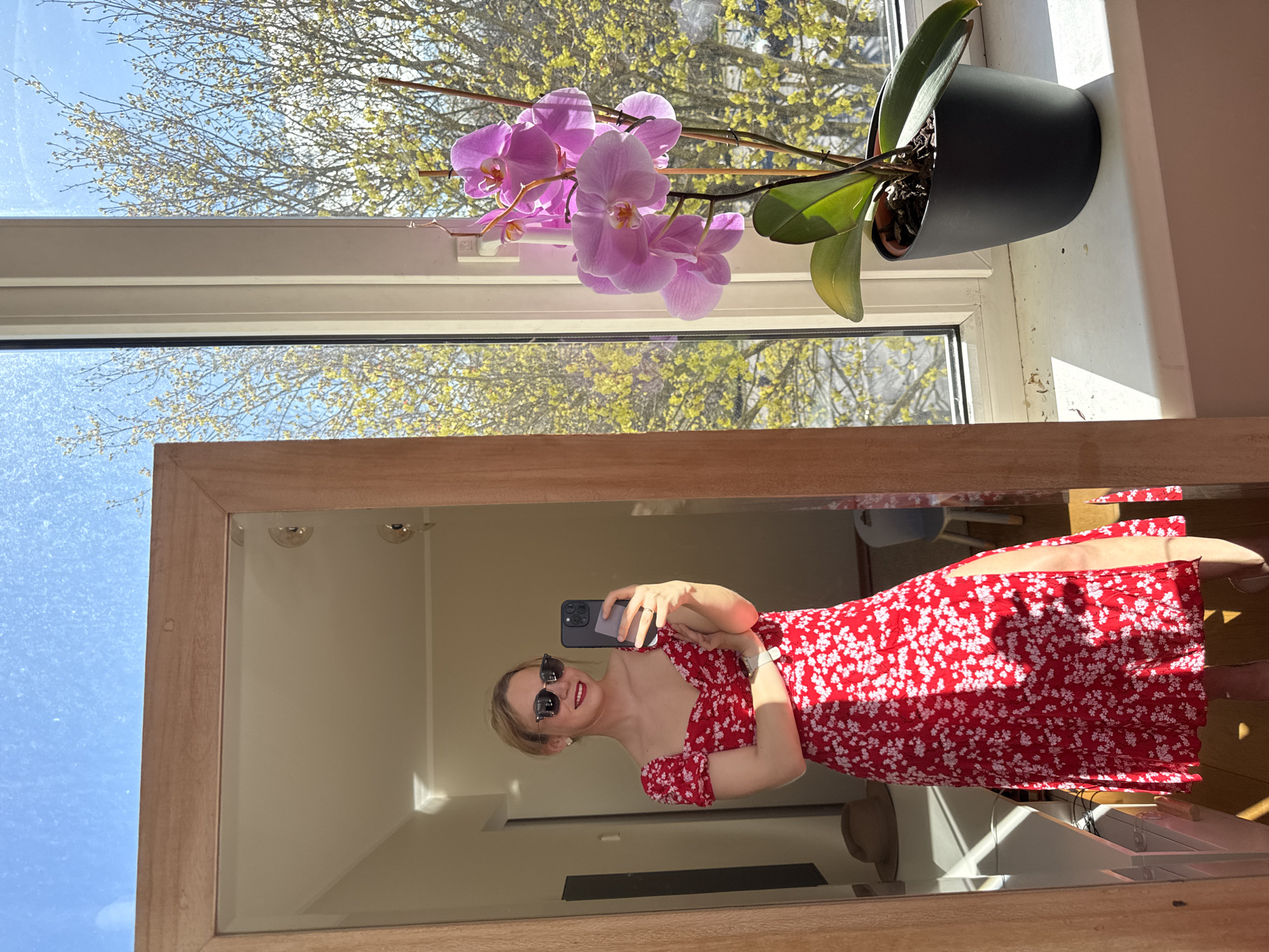 This image captures a mirror selfie taken by a young woman in an indoor setting, with a bright outdoor view through a window.
In the foreground, a large, rectangular mirror with a light wooden frame reflects the woman. She is fair-skinned, with blonde hair pulled back, and wears black sunglasses and red lipstick, sporting a visible smile. She is dressed in a vibrant red dress with small white floral patterns and an off-the-shoulder, ruched neckline. Her right hand holds a dark smartphone, used to capture the selfie, while a ring is visible on her left hand and a watch on her left wrist.
To the right of the mirror, a windowsill holds a potted orchid with several delicate pink-purple flowers and green leaves in a dark grey pot. Behind the orchid, a window reveals a sunny outdoor scene. The sky is bright blue, and trees with young, light green, budding leaves are visible, strongly suggesting springtime. The sunlight streams into the room, illuminating the orchid and reflecting brightly off surfaces.
The activity taking place is a mirror selfie, capturing both the individual and a glimpse of their environment. The bright natural light indicates it is daytime, likely a clear and pleasant day. The budding trees specifically point to the spring season. The woman's smile conveys a sense of happiness or contentment. Based on the provided context, the location is Riga, Latvia. No visible text is present in the image.