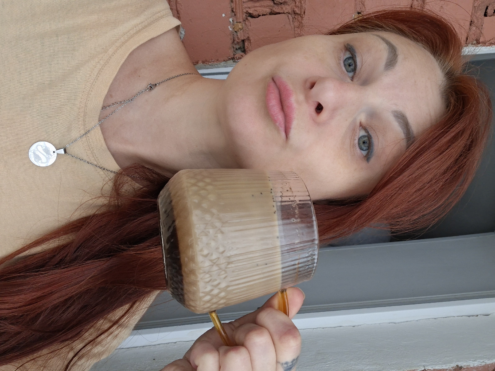 A woman with long, auburn hair and pale skin is holding a clear, ribbed mug filled with a light brown beverage, likely coffee, in her right hand. She is wearing a light yellow t-shirt and a silver necklace with a circular pendant. The mug has an amber-colored handle. Her head is tilted slightly, and she is looking towards the camera with a neutral expression. The background includes a red brick wall on the left and a white door frame on the right, suggesting an outdoor or semi-outdoor setting. The scene appears to be a close-up portrait. There are no identifiable landmarks or features that would specifically place this image in Myadzyel, Belarus.