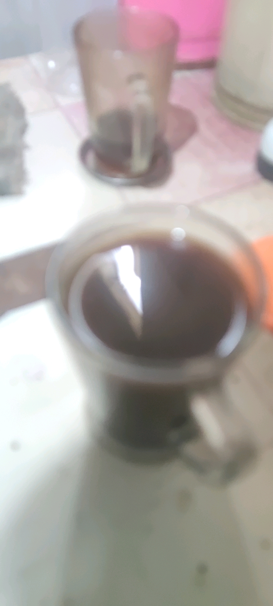 The image is a close-up, out-of-focus shot of a glass mug filled with a dark liquid, likely coffee, with a bright reflection on the surface. The mug has a clear handle. In the background, there are other blurred objects, including what appears to be another glass or container and a pink object. The setting seems to be a table with a textured surface, possibly a tiled countertop or placemat. There is no indication of the specific location within Gambarsari, Indonesia, or any details about people or actions taking place.