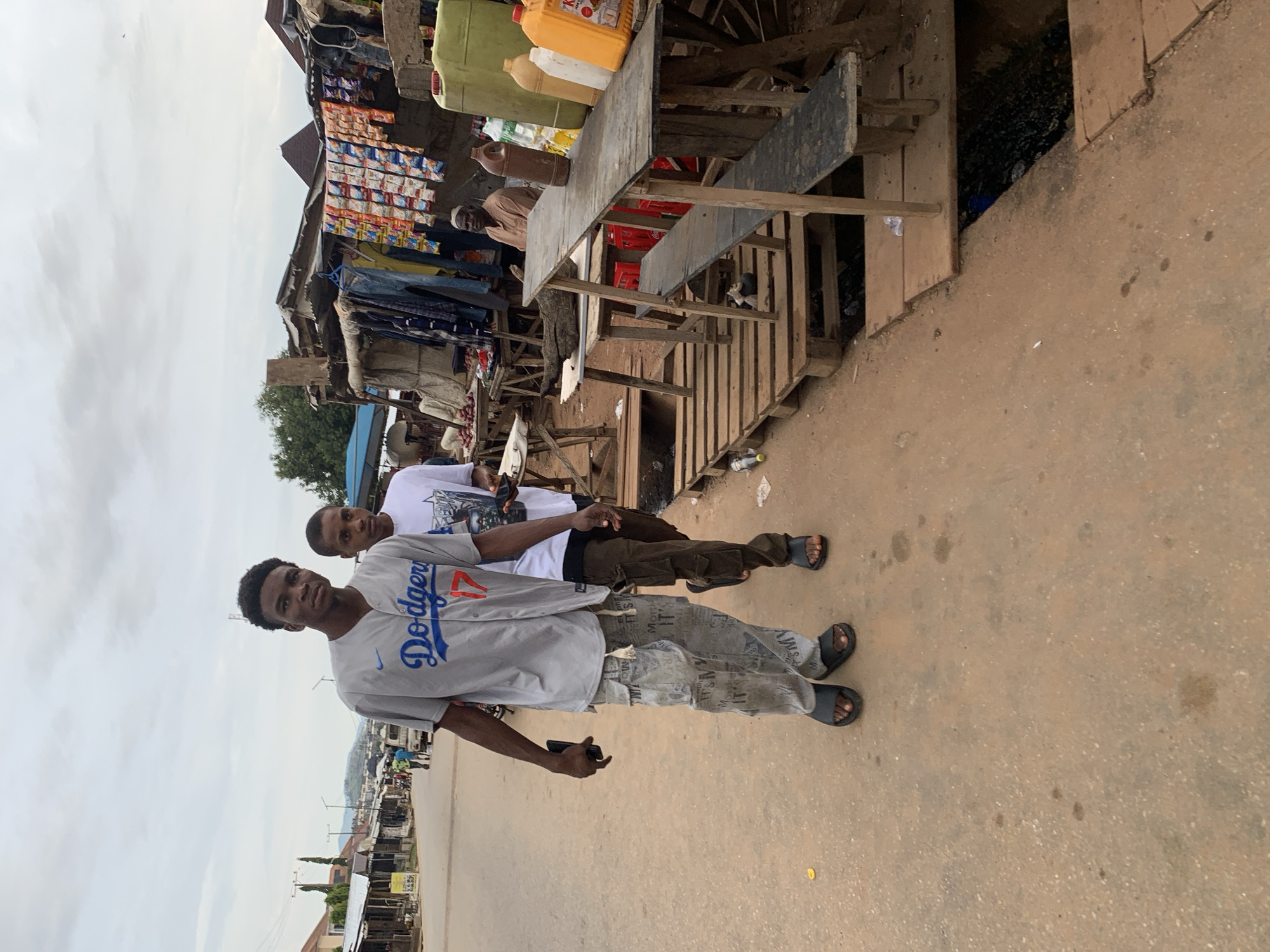 This image depicts a street scene in Abuja, Nigeria. Two young men are walking on a paved road with a makeshift market stall on the right side. The man in the foreground is wearing a grey baseball jersey with "Dodgers" and the number "17" on it, paired with baggy, printed pants and black sandals. He is holding a dark-colored phone in his left hand. The second man, slightly behind and to his right, is wearing a white t-shirt and dark cargo pants with black sandals. He is carrying a white plate with food.
The market stall features a rustic setup with wooden planks and corrugated metal roofing. Various items are displayed, including clothing hung from a rack, packaged goods arranged in rows, and several large plastic containers of liquid. A man in a light brown shirt is seated behind the stall. The environment suggests a typical bustling street market with an open drainage channel visible in front of the stall, covered in part by wooden planks. The sky is overcast, indicating a cloudy day. The overall impression is one of a lively, informal commercial area within the city.