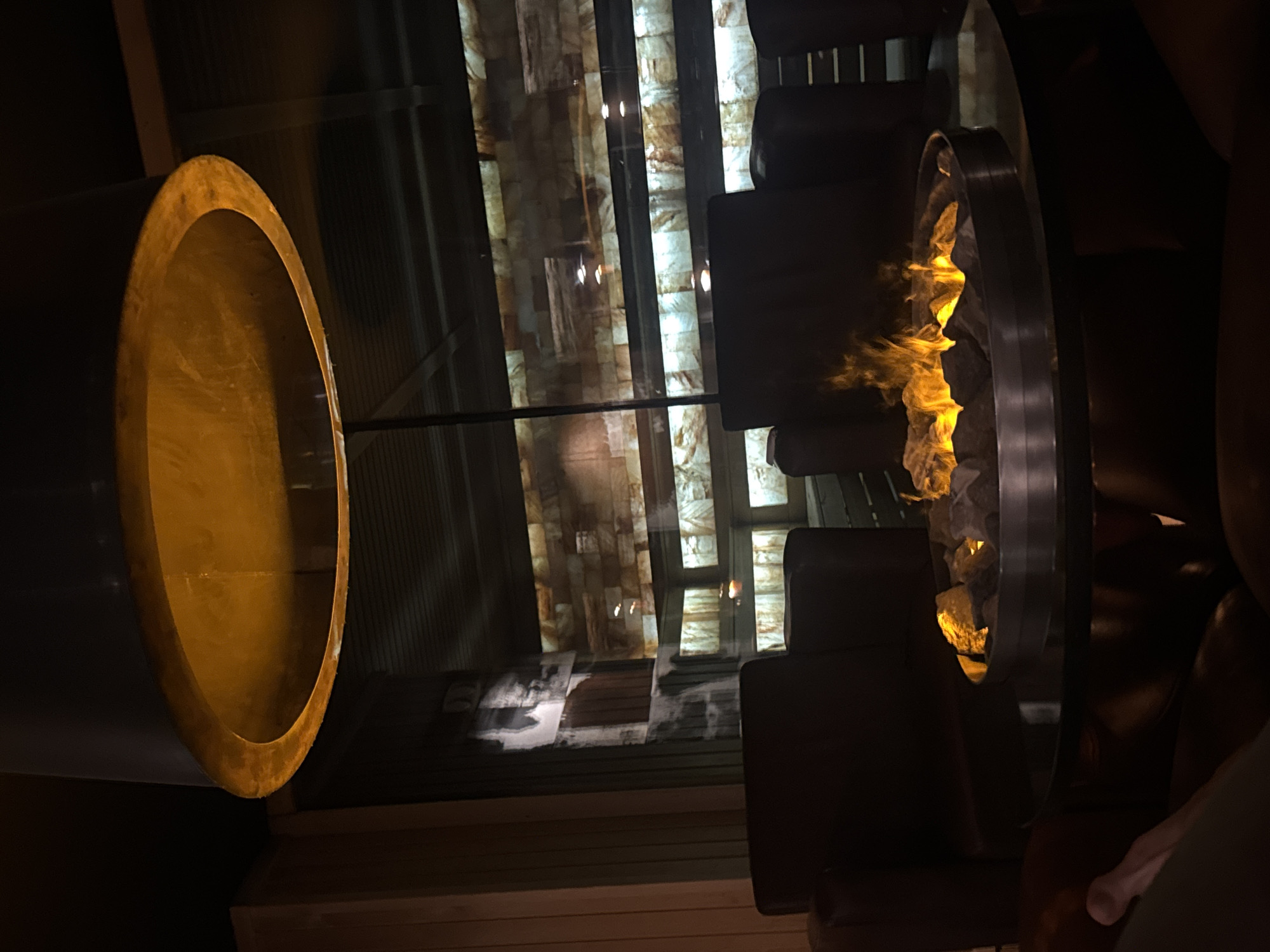 The image depicts an indoor lounge or bar environment characterized by dim, warm lighting. A prominent circular fire pit table, featuring a metal ring filled with stones from which orange-yellow flames emanate, occupies the center foreground. Dark, presumably leather, armchairs are arranged around this table. A human leg, partially visible in the lower left, indicates the presence of an occupant. Directly overhead, a large, inverted, dark drum-shaped light fixture with a reflective, warm-toned interior casts a golden glow. The background features a large glass partition, behind which a wall composed of horizontally laid, backlit translucent blocks (possibly onyx or salt) creates a glowing, textured effect. Vertical structural elements are visible through the glass. To the far left, a section of wooden paneling is visible. The scene suggests a cozy and modern establishment. This location is identified as Infinity Rooftop Plovdiv, situated within the Grand Hotel Plovdiv in Plovdiv, Bulgaria.