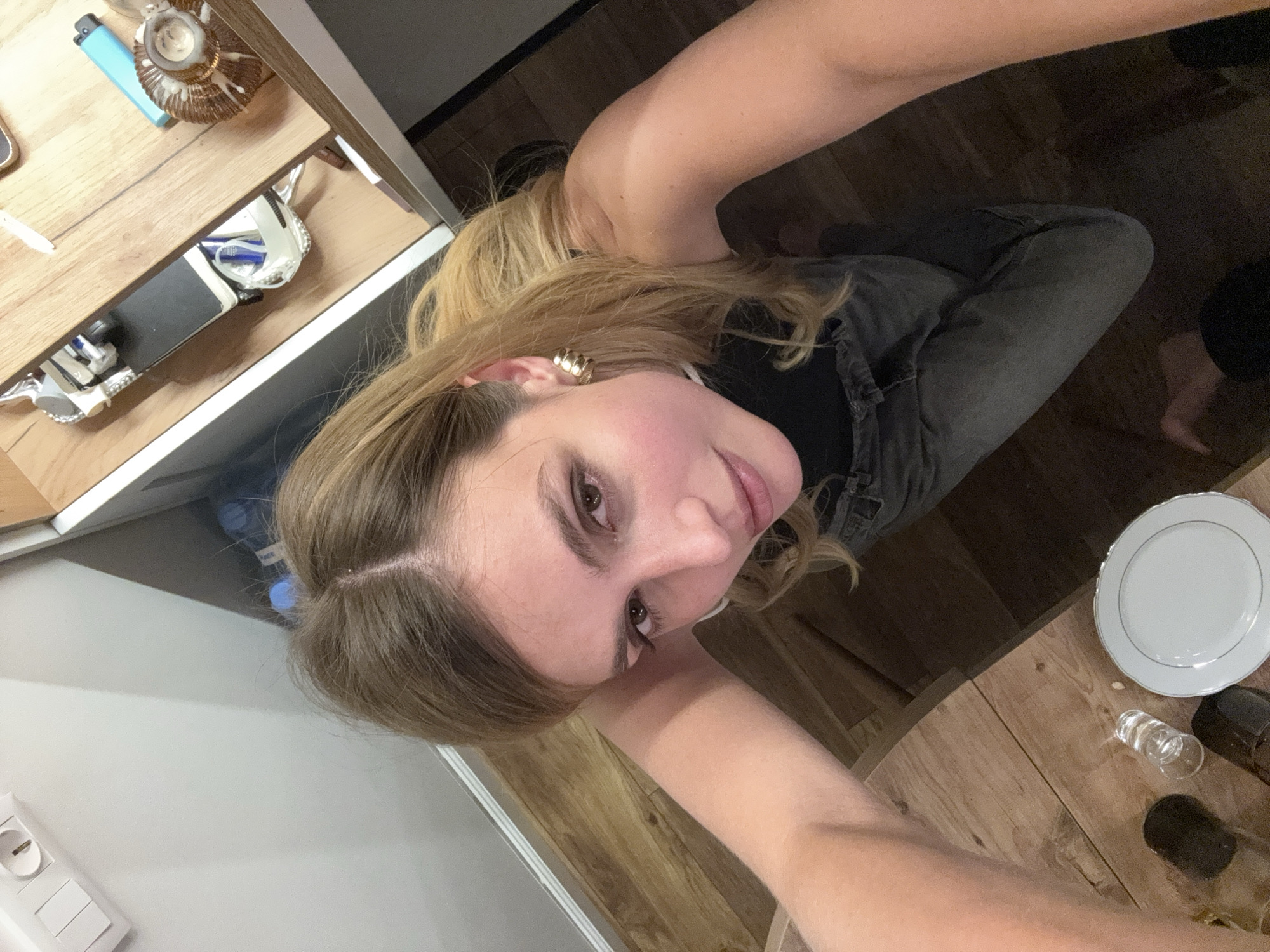A top-down selfie of a blonde woman with fair skin, wearing a gold hoop earring and a black top with gray jeans, is taken in what appears to be a residential interior.
The background includes wood flooring and what may be a wooden dining table with a white plate and a drinking glass upon it. To the left, there is a light gray or white cabinet or countertop with items on top including a lighter, a small decorative shell, and other smaller unknown objects.
The location is Unknown, Unknown.