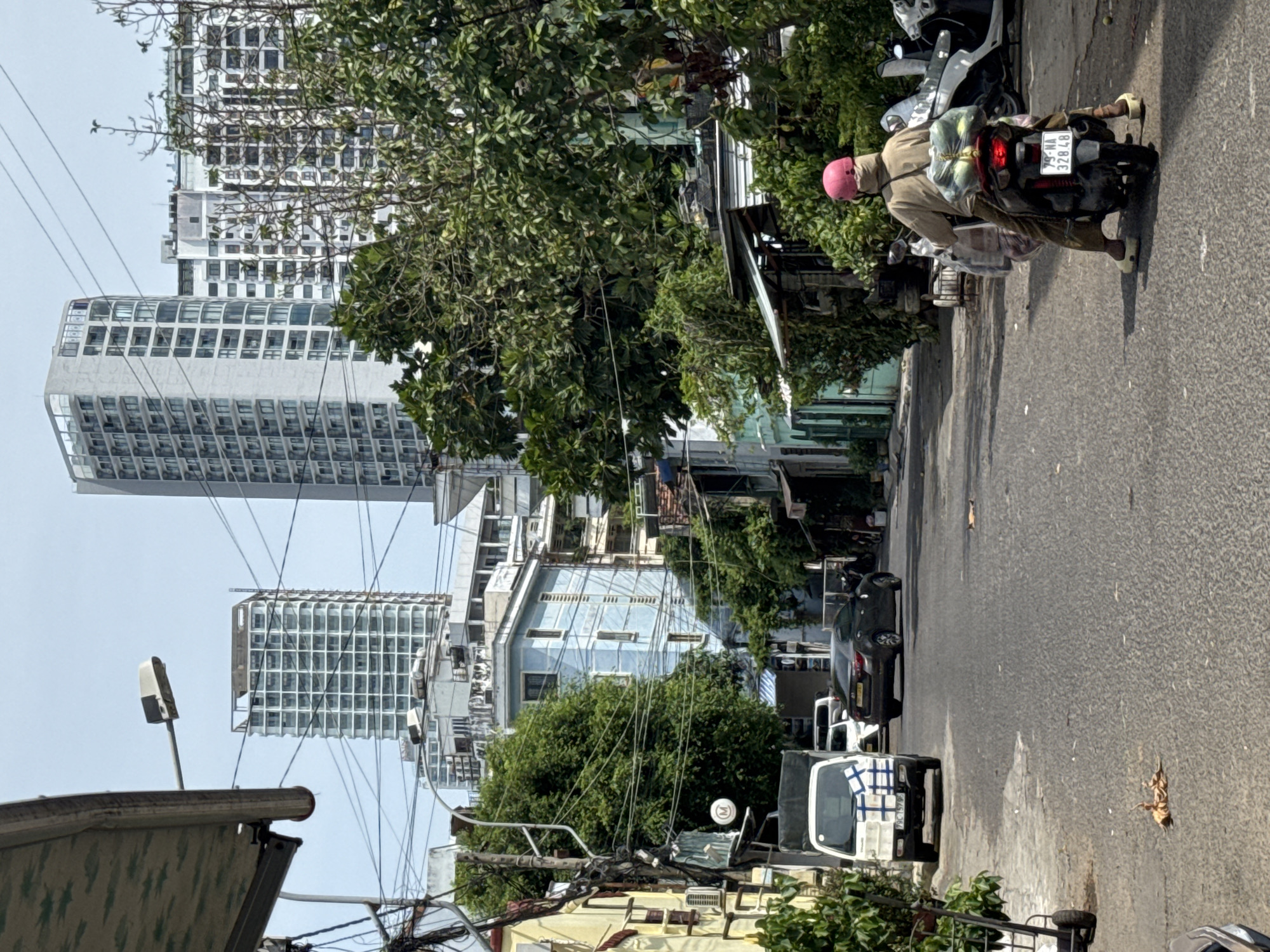 In a city street scene in Nha Trang Ward, Vietnam, a person wearing a light brown hooded garment and a pink helmet rides a dark-colored scooter. The scooter is loaded with multiple bags, including what appears to be produce and other items strapped to the back. The rider's feet are clad in light-colored footwear. They are on an asphalt road with some scattered debris. To the left, a white delivery van with blue markings on its side is parked. Further down the road, a dark sedan is visible. The street is lined with various buildings, including older structures and taller, modern high-rises in the background. Lush green trees and foliage are abundant, with dense branches obscuring parts of the buildings. Overhead, a street lamp is mounted on a pole, and numerous electrical wires crisscross the scene. The sky is clear and bright.